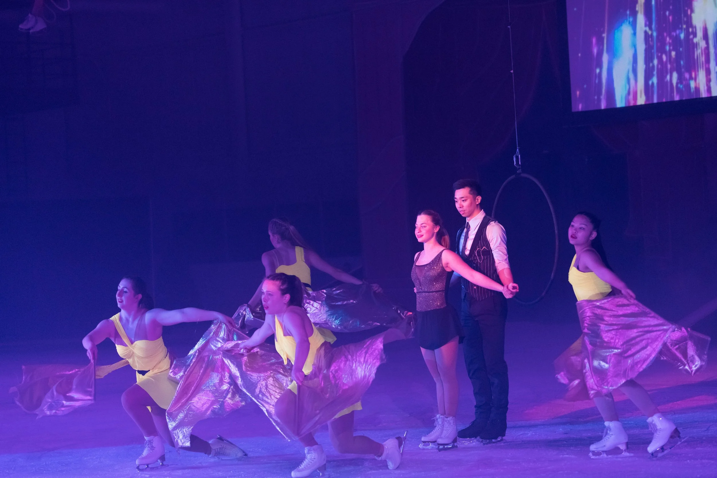 A group of young dancers performing on stage under purple lighting, dressed in colorful costumes, with some holding metallic fabric props.