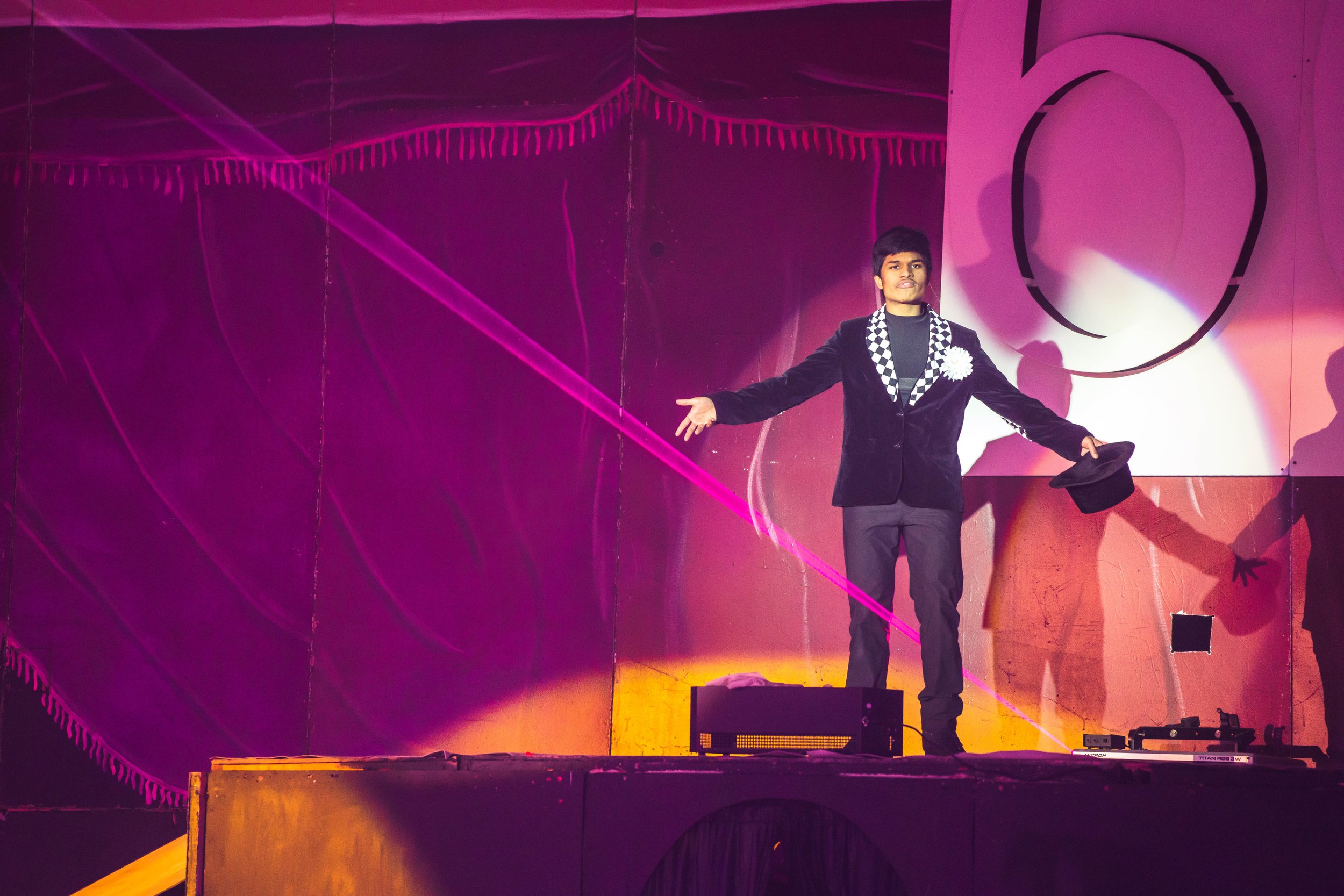 A man in a black blazer with a checkered collar and a large white flower pinned to his chest stands with arms open on a colorful stage with pink and purple lights, a mirror, and a large black circle in the background.