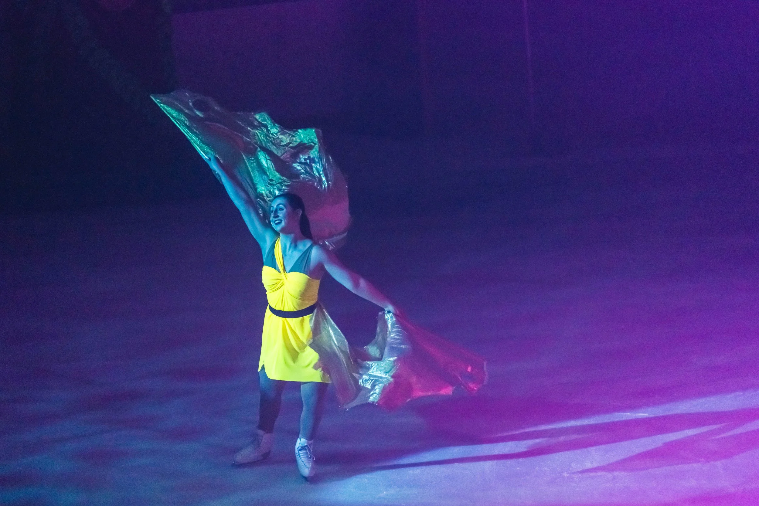 A female dancer performing under colorful stage lights, wearing a yellow dress with black accents, holding a metallic fabric in her hand, with a joyful expression.