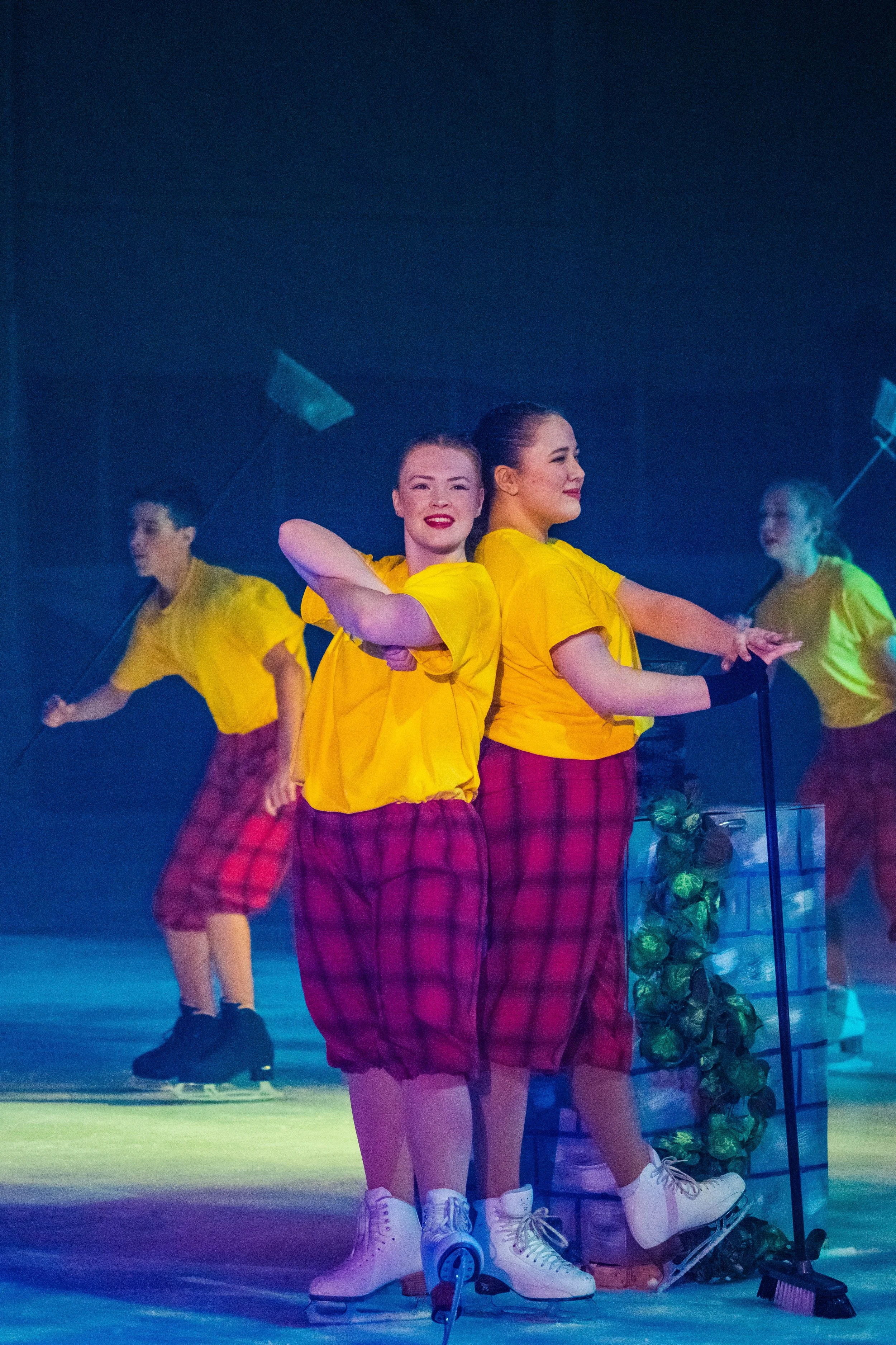 Four women in yellow shirts and red plaid skirts ice skating on a stage with a blue background, a mirror, and a decorative stand with flowers.