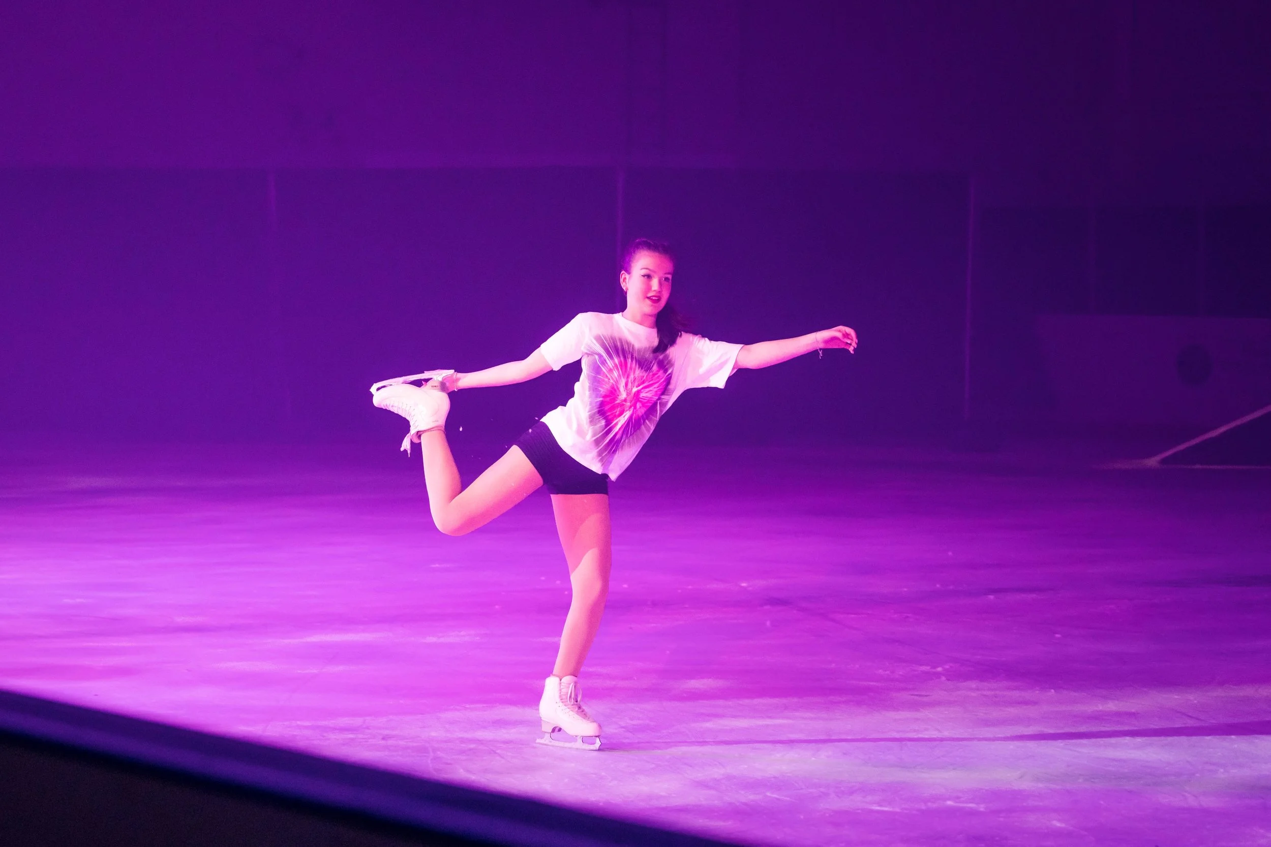 A young woman figure skater is performing a balanced pose on ice, wearing a casual t-shirt and black shorts, with vibrant purple and pink stage lighting in the background.