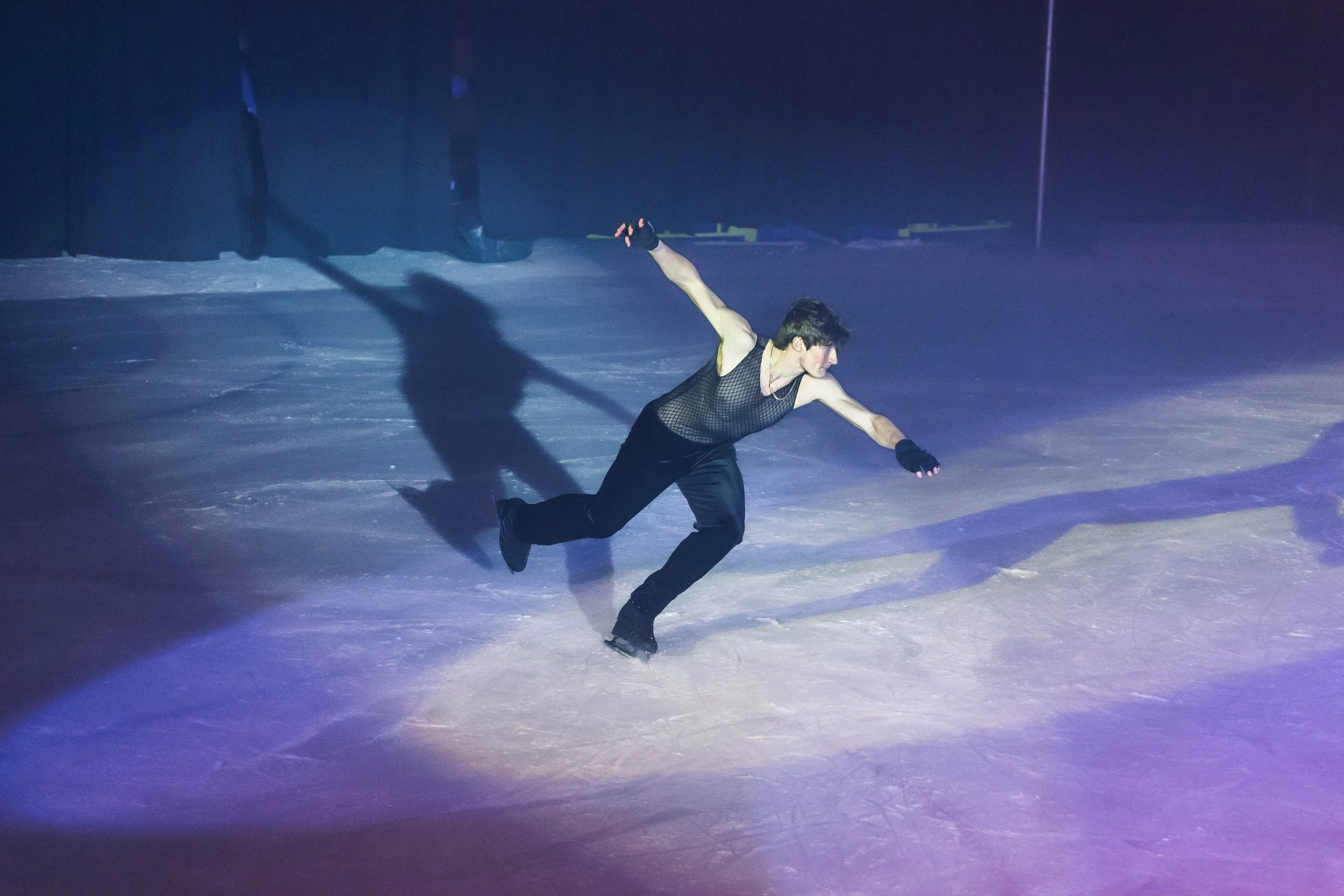 A male figure skater wearing a black mesh top and black pants glides across the ice rink with one arm extended and the other bent behind him, in a pose during a performance or practice.