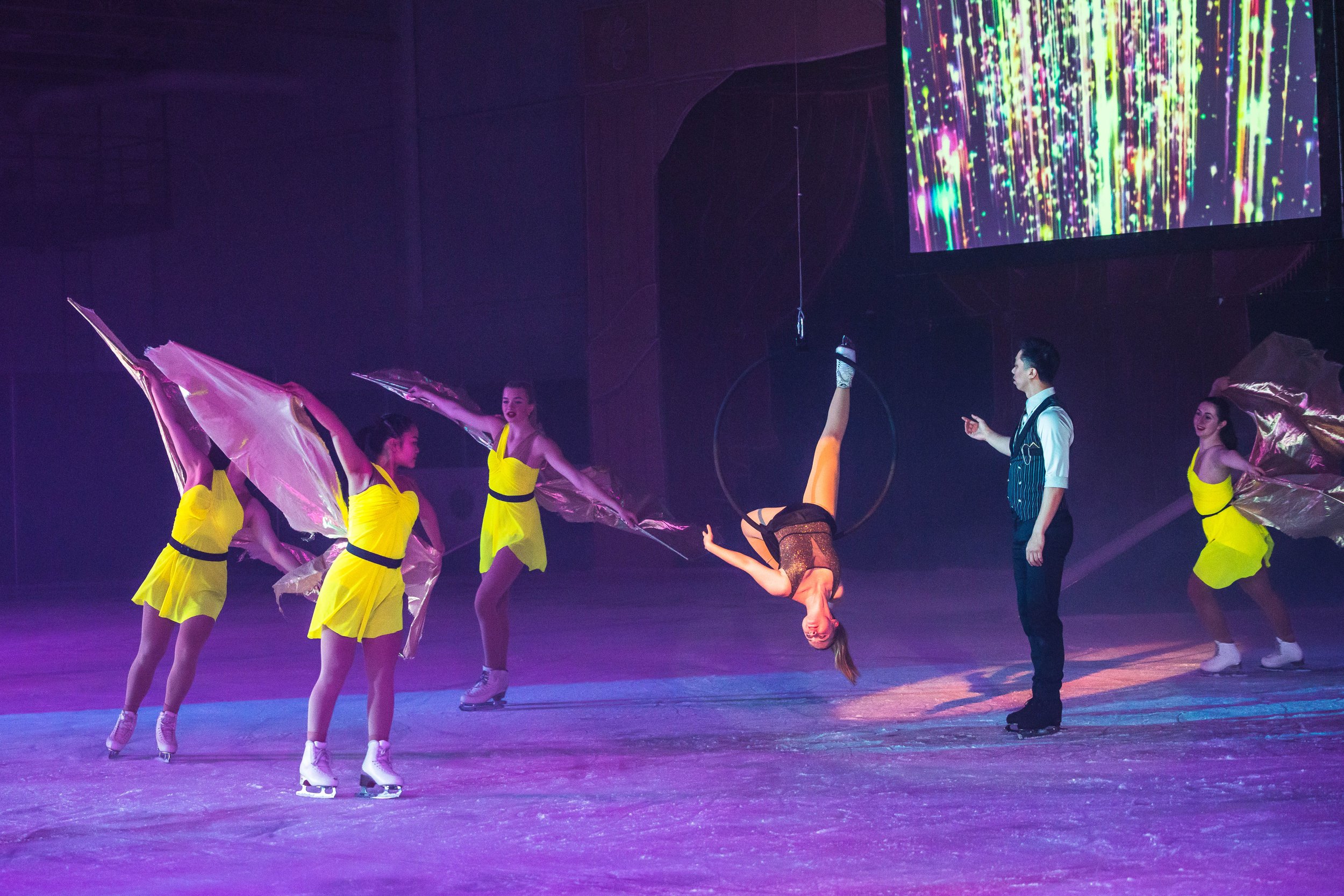 Figure skaters in yellow costumes performing a routine on ice, with one dancer upside down, in front of an audience in a darkened theater with colorful digital display on a large screen.