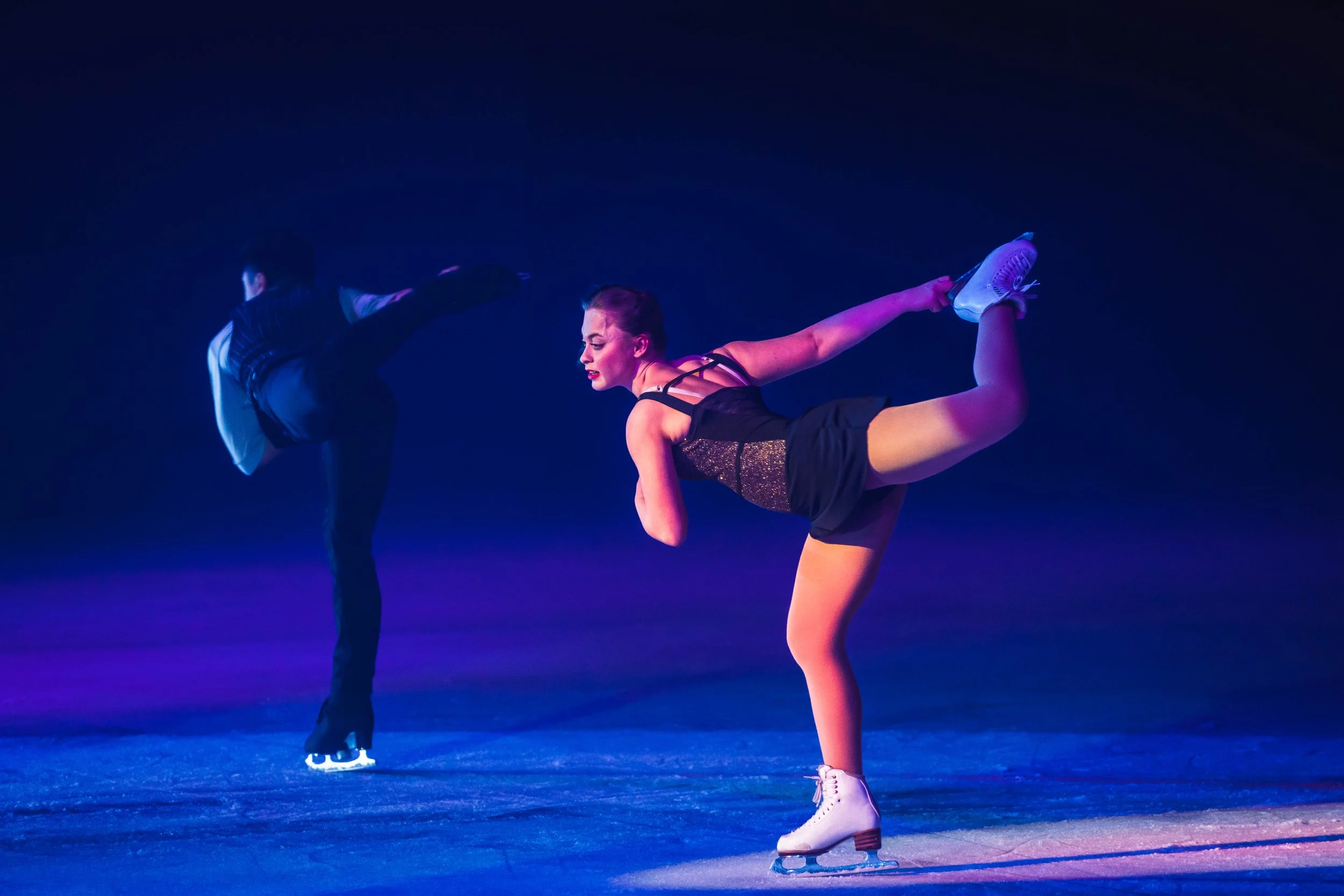 A female figure skater performing a dance move on ice, extending her leg back while holding her skate with one hand, wearing a black dress with sparkles and white ice skates. A male figure skater is partially visible in the background, dressed in a d