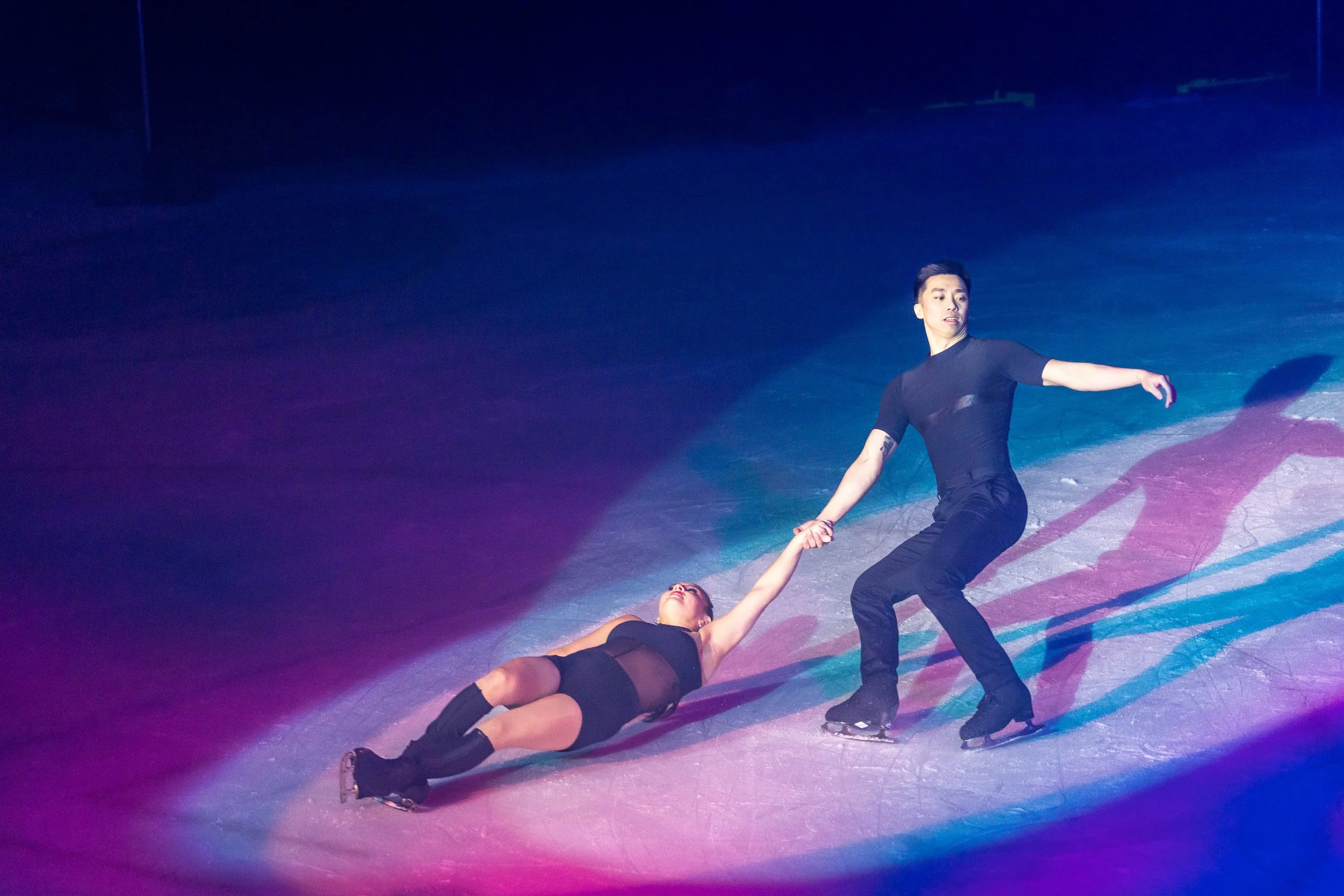 Two figure skaters are performing on the ice, with one lying on her back and the other standing, holding her hand, in a dark, colorful setting.
