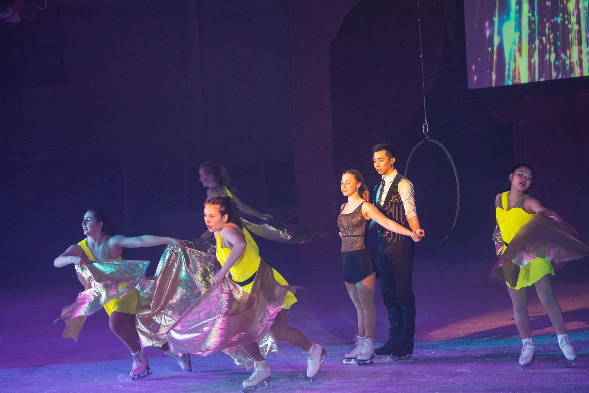 Group of young performers dancing on stage with costumes featuring shiny yellow fabric and ice skates, under stage lighting.