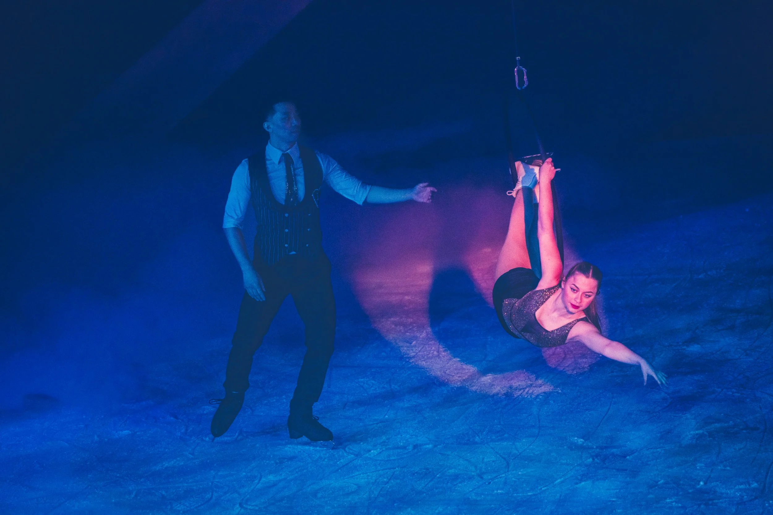 A male performer with a vest and tie, holding a pole with a suspended object, and a female performer in a glittery dress, performing a dance move on a stage with blue and purple lighting.