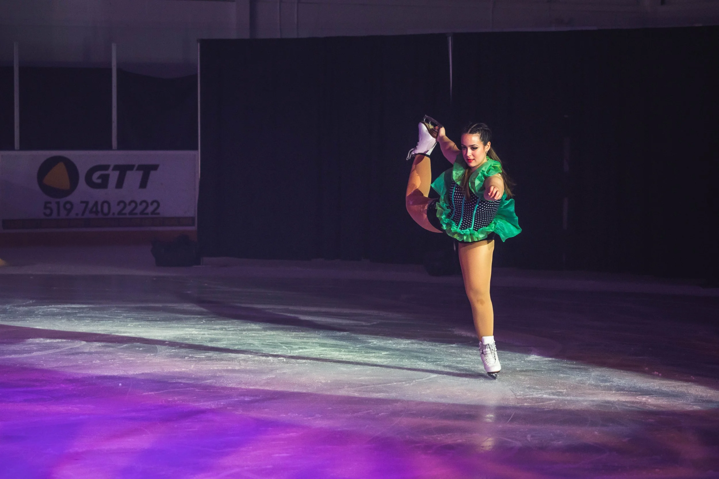 A female figure skater performing a spin on ice, wearing a colorful costume with green and black, in an ice rink.
