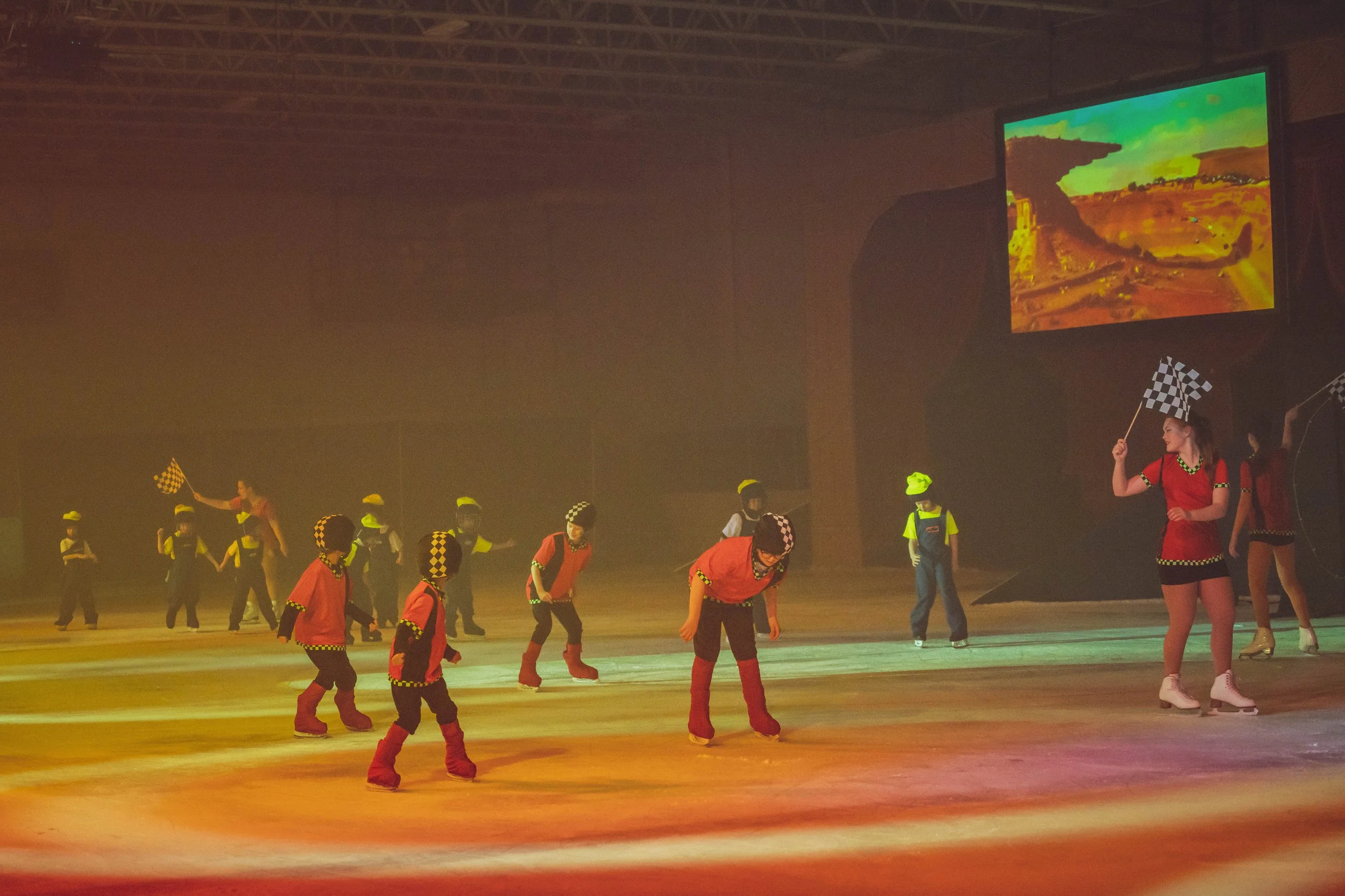 Group of children in colorful costumes performing on stage with two women, one holding a checkered flag, under colorful stage lighting.