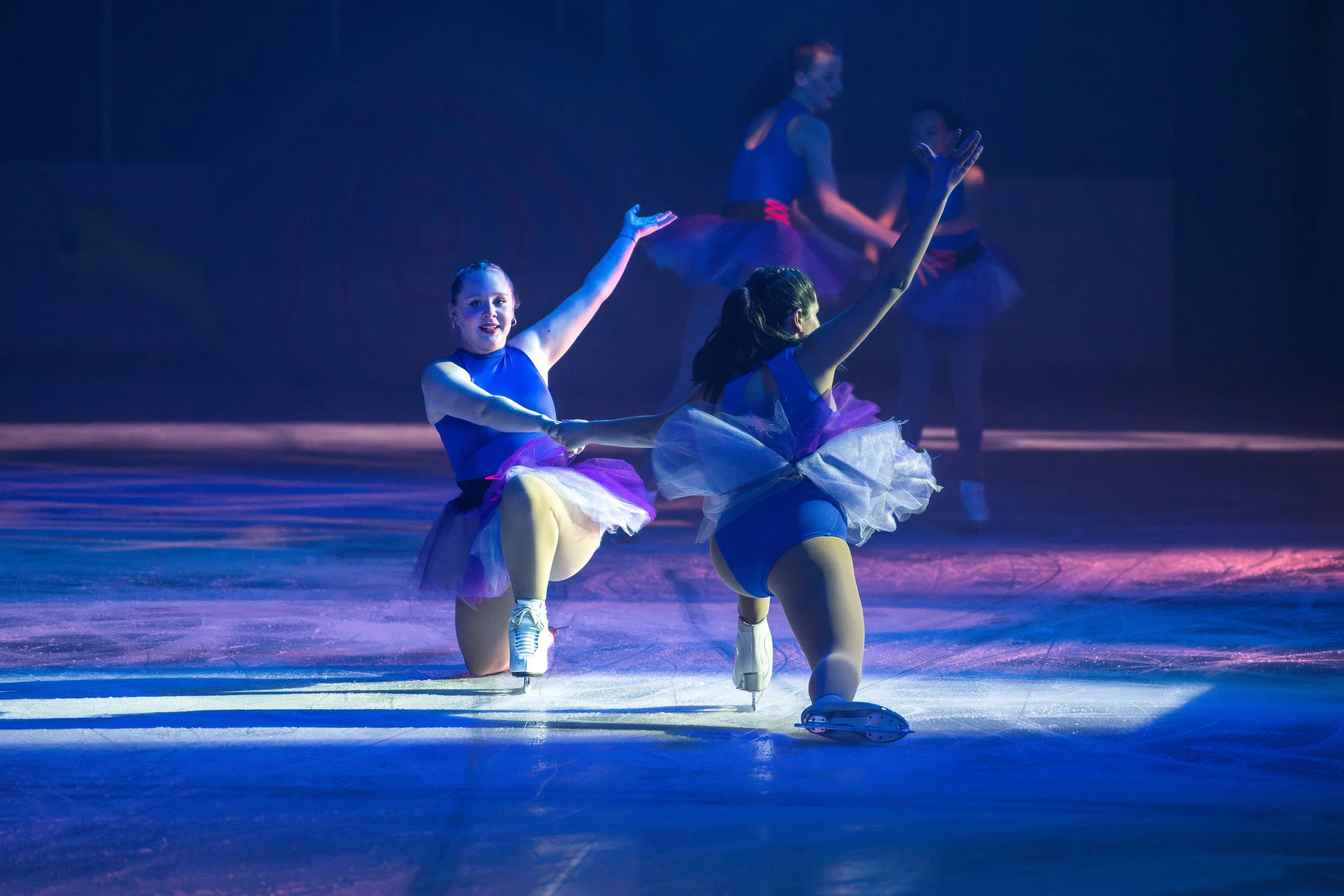 Two young female figure skaters dressed in blue costumes performing a routine on the ice, with one kneeling and the other standing, in a dark setting with blue and purple lighting.