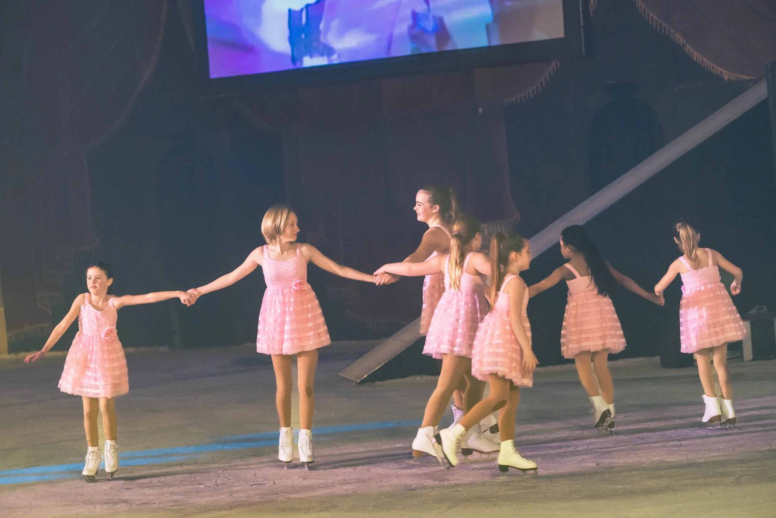 A group of young girls in pink plaid dresses ice skating in a line, holding hands, in a large indoor skating rink with a large screen above them.