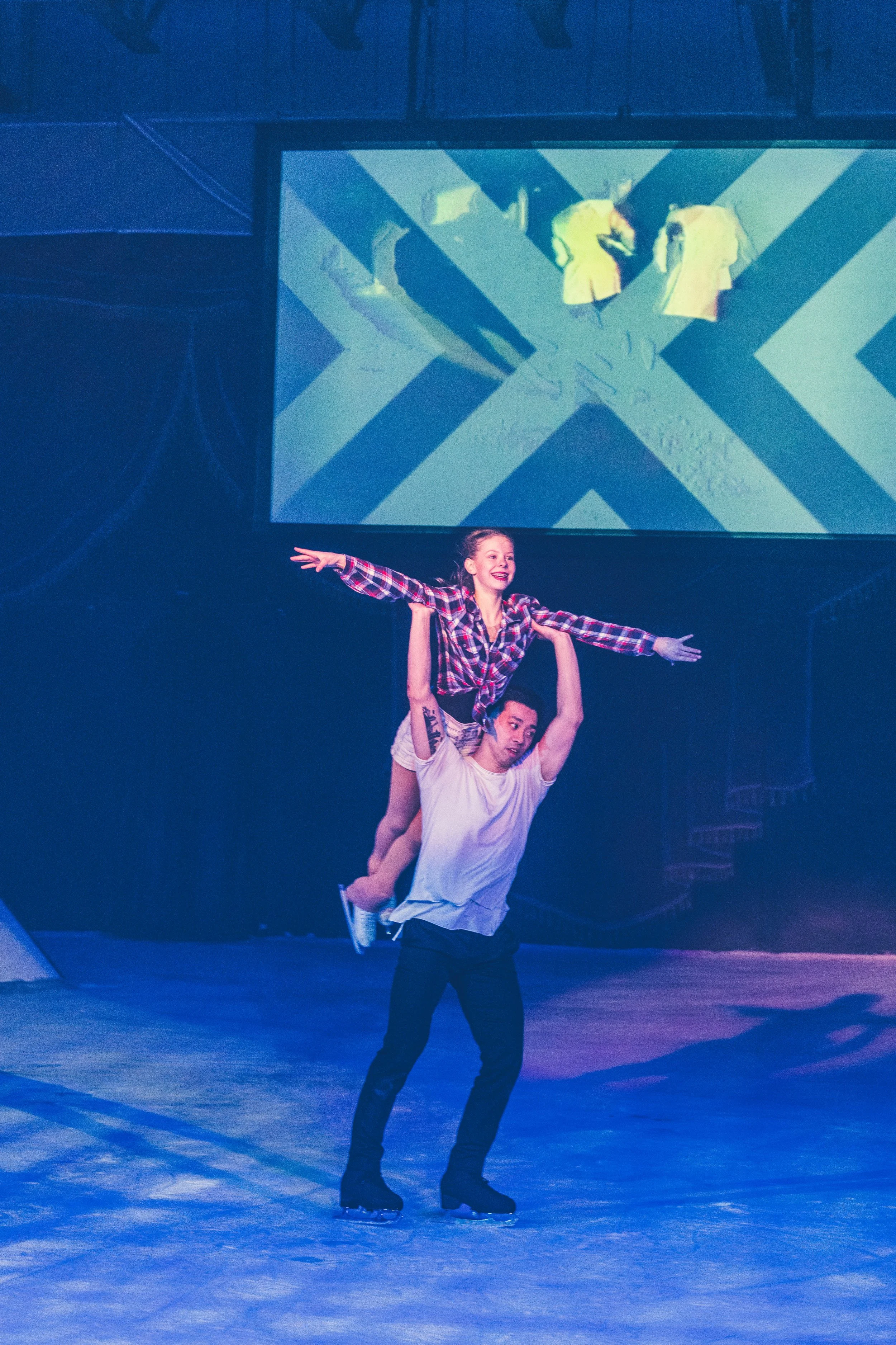 Two ice skaters perform a routine at an indoor ice rink. One skater, standing on the ice, is seen holding the legs of the other skater, who is balanced on her hands with her arms extended outward. The background features a large screen displaying a g