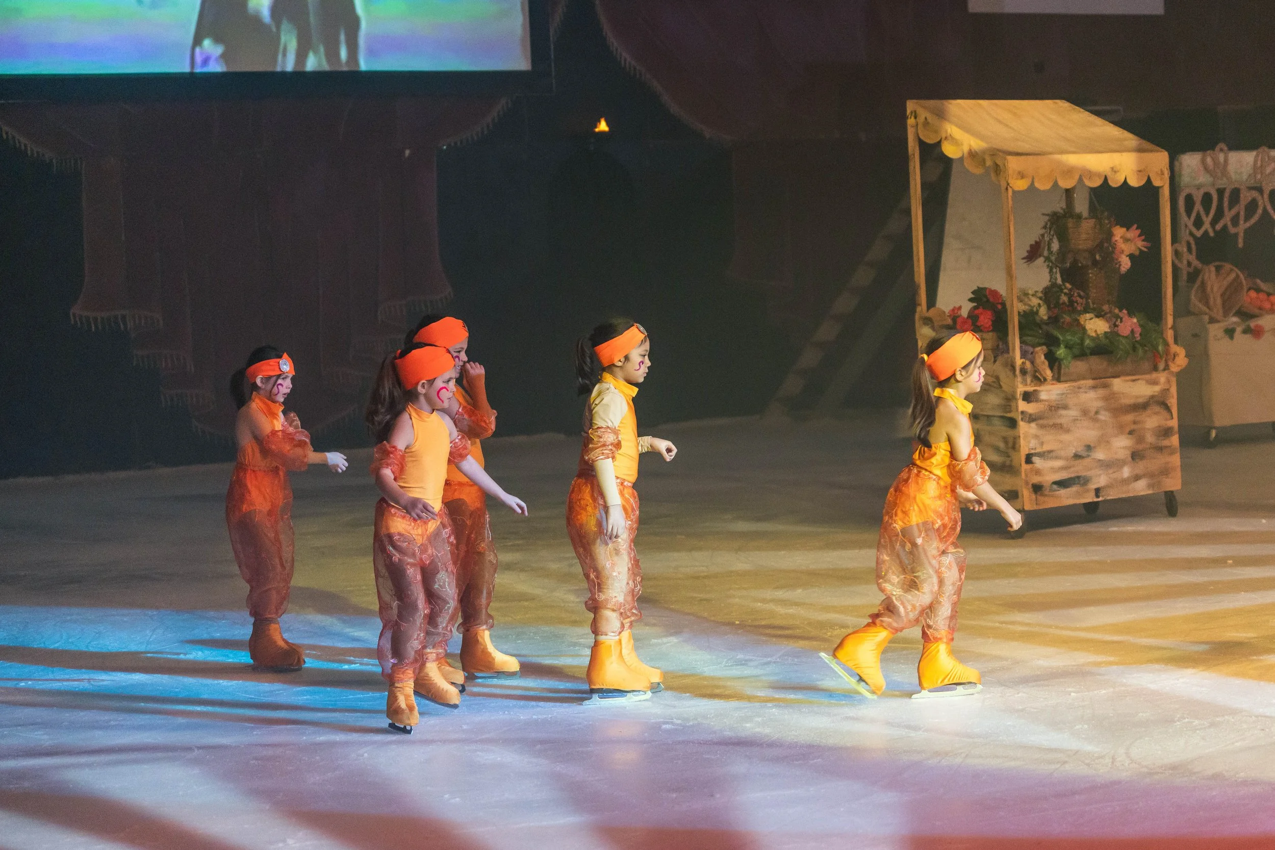 Young children dressed in orange costumes ice skating in an indoor rink, with a decorated cart and a large screen showing a performance in the background.