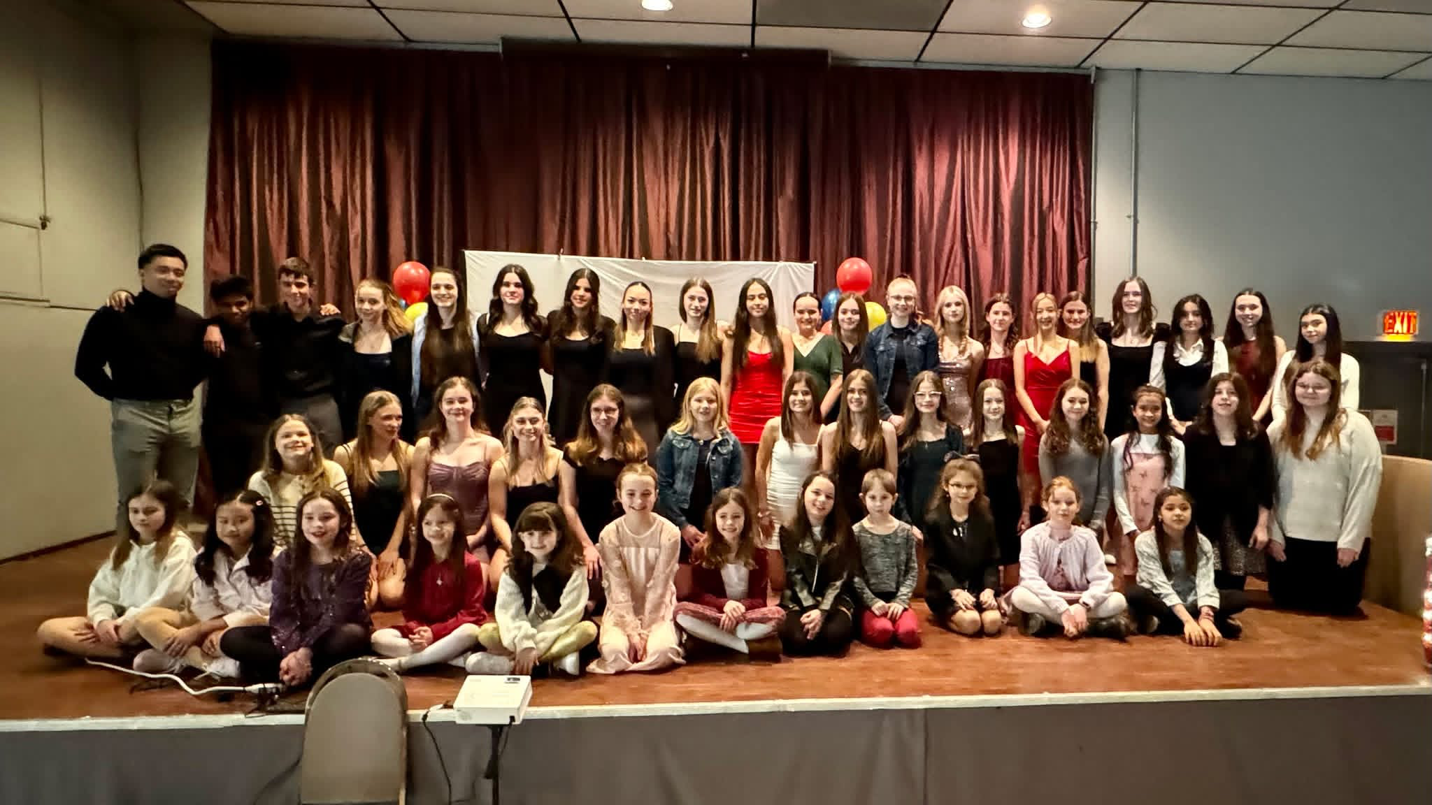 A large group of people, including children and adults, gathered together on a stage in front of a red curtain, posing for a group photo.