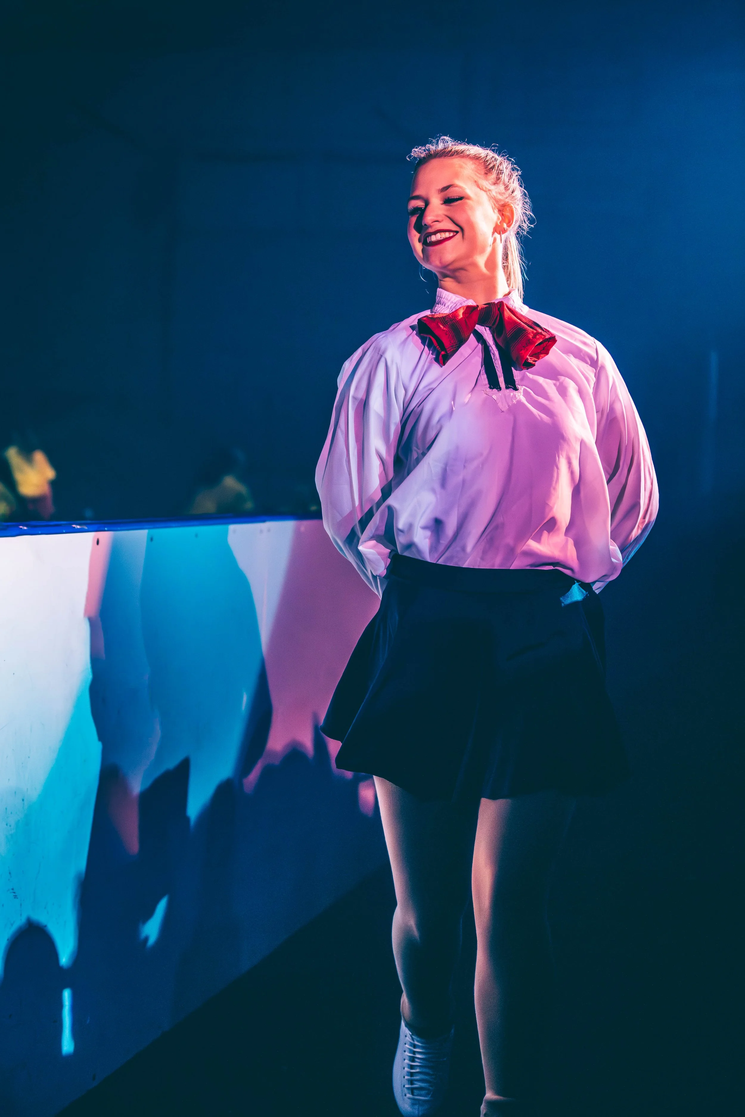 A young woman with blonde hair tied back, smiling, wearing a white shirt with a red plaid bow tie, a black skirt, and white sneakers, standing by a wall with colorful reflections.