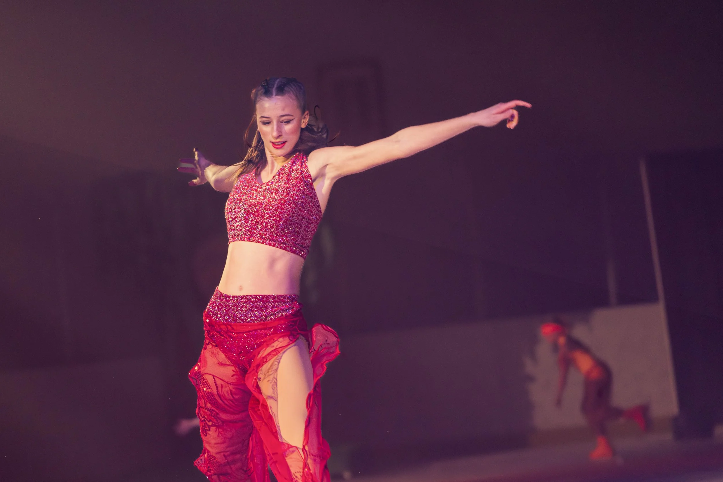 A female dancer in a red, patterned costume performing on stage with her arms extended outward.