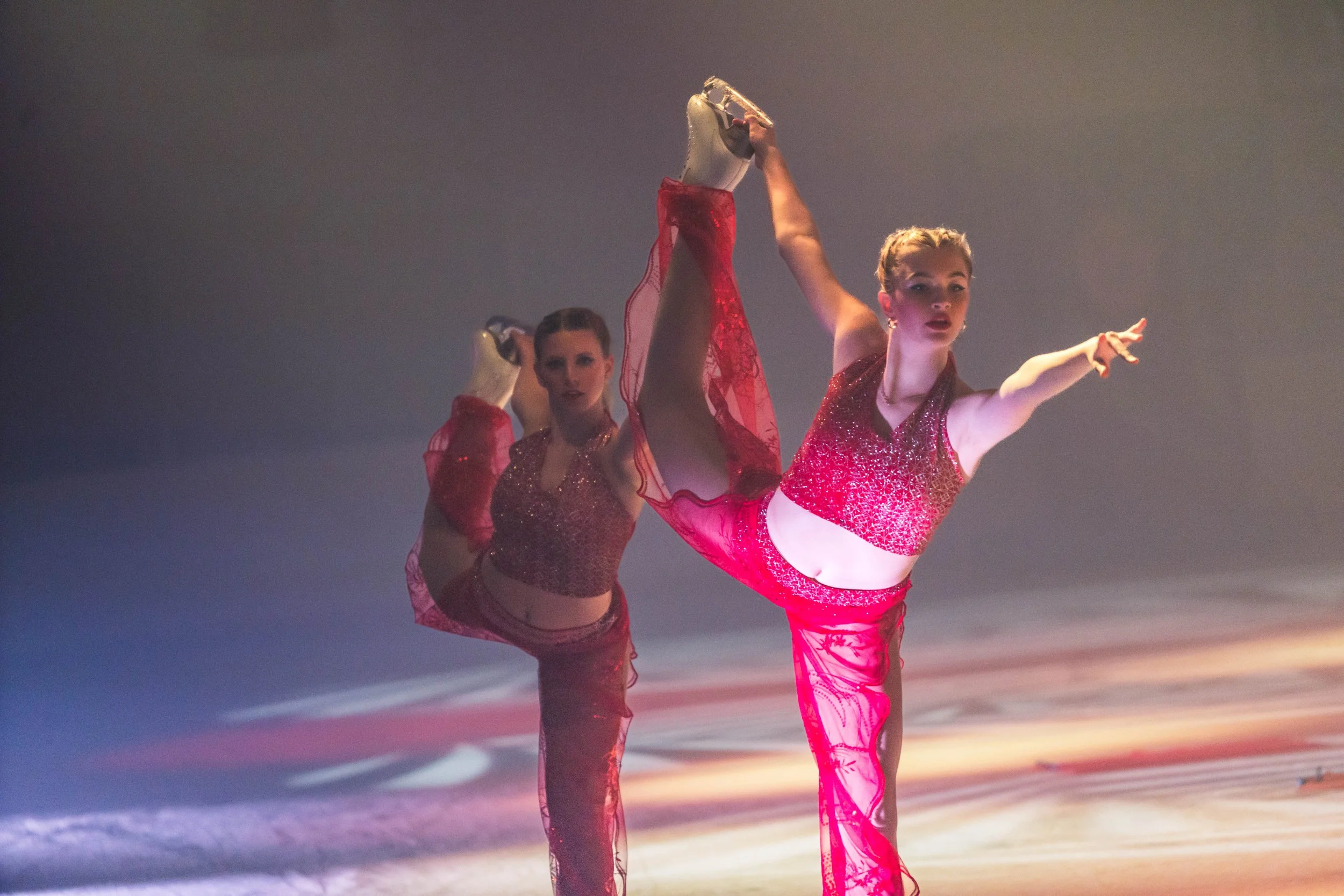 Two female ice skaters in red costumes performing a synchronized routine on the ice; one skater is executing a move with her leg extended high and holding her skate above her head, the other skater is behind her on the ice.