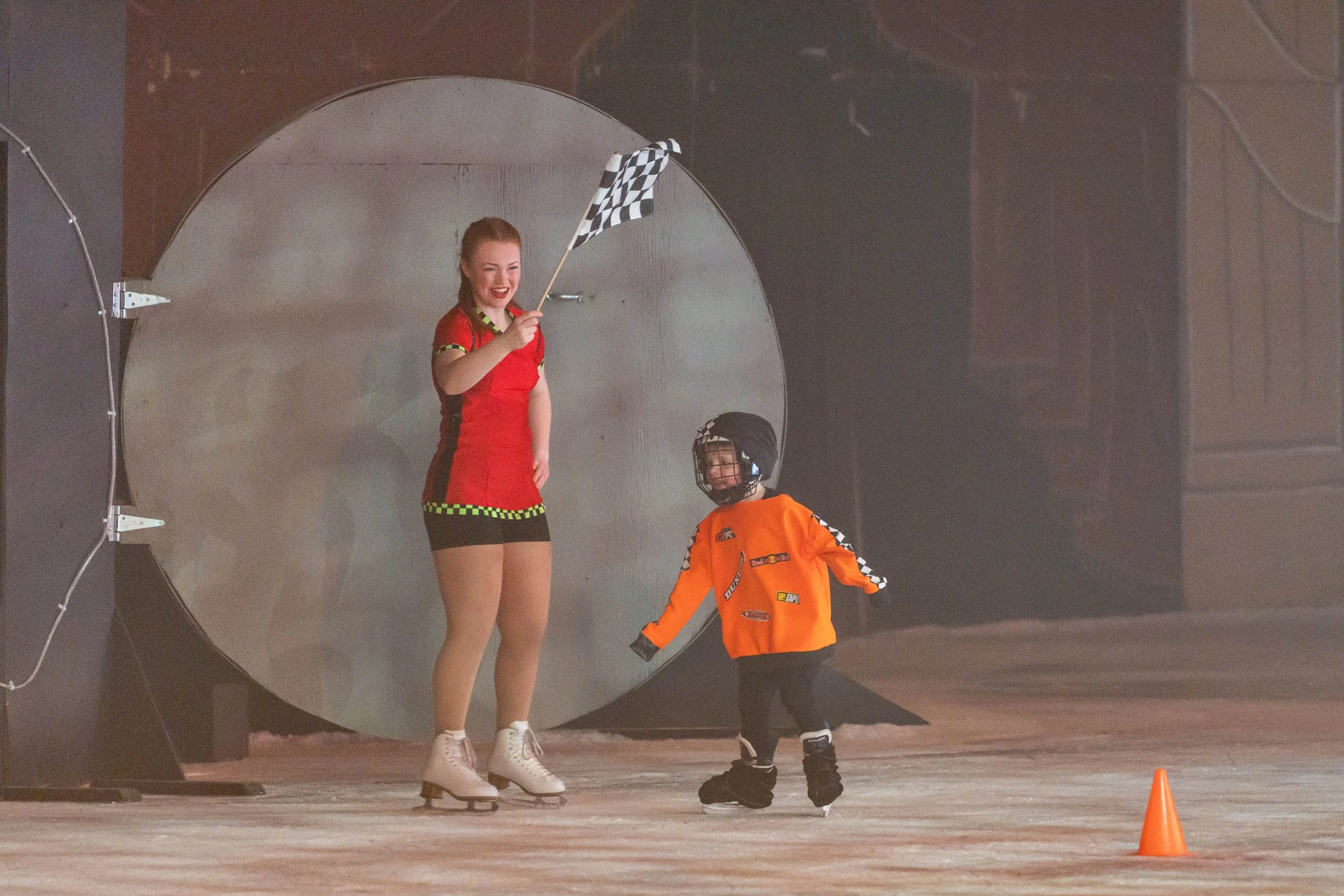 A young woman in a red shirt and black shorts with a checkered trim is standing on ice skates, holding a checkered flag, smiling. A small child in an orange jacket, black helmet, and black skates reaches out to her, with an orange cone visible on the