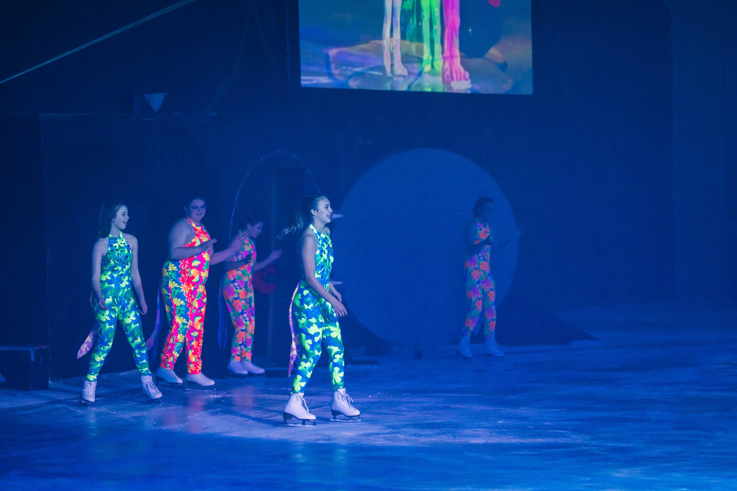 Group of five women in colorful floral dresses performing on stage with ice skates, under blue lighting, with a large screen displaying their legs in the background.