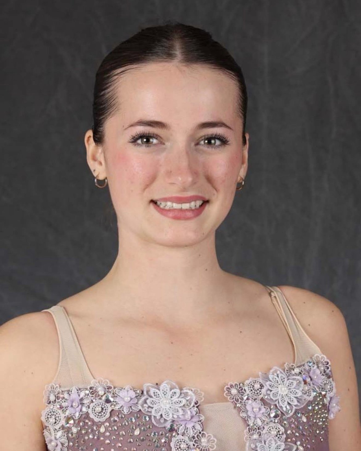 A young woman with dark brown hair, tied back, wearing a sleeveless dress with floral embroidery and rhinestones, smiling at the camera against a dark background.