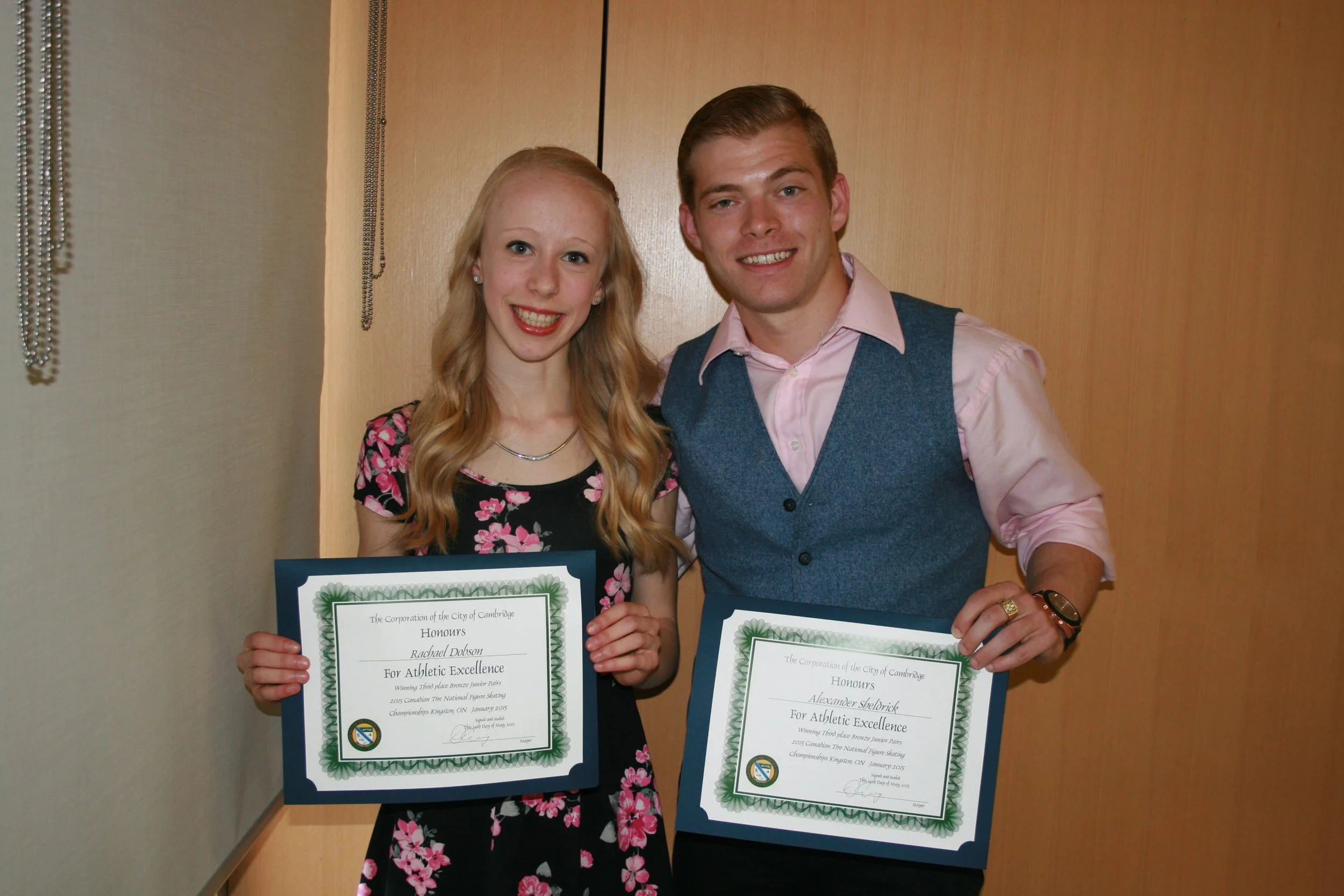 Two young adults, a woman and a man, smiling and holding certificates of achievement, standing together indoors against a wooden wall.