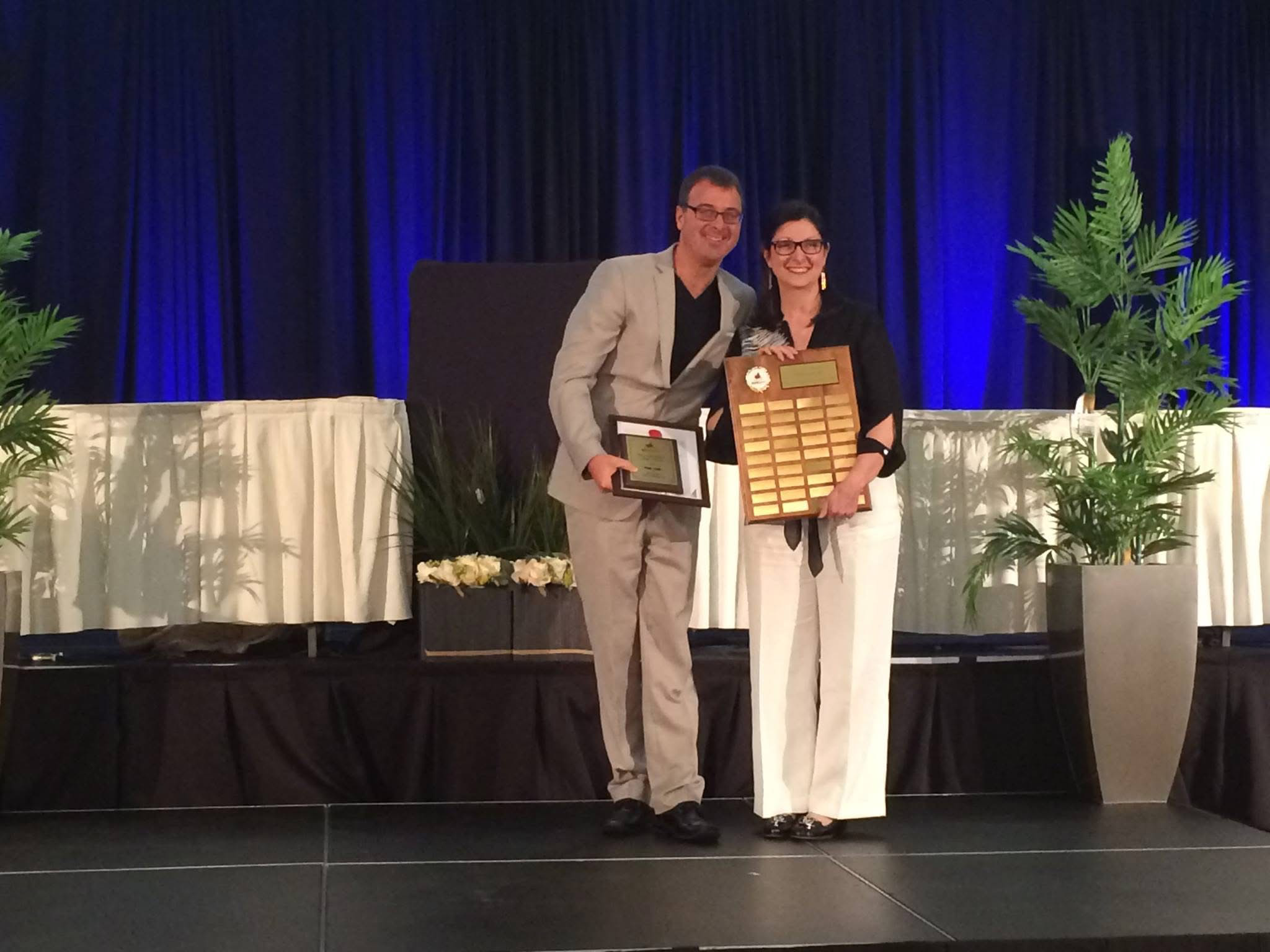 Two people on a stage holding plaques and smiling, with a blue curtain background and potted plants on either side.