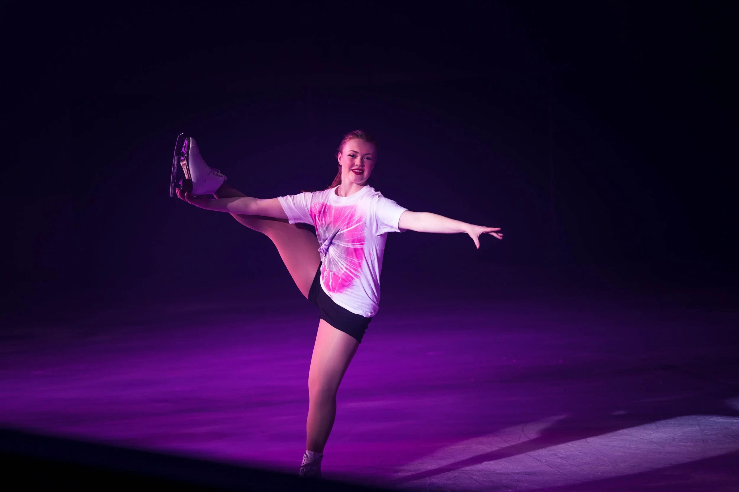 A young female figure skater performing a high kick on ice, wearing a white T-shirt with pink design and black shorts, with a dark background and purple stage lighting.