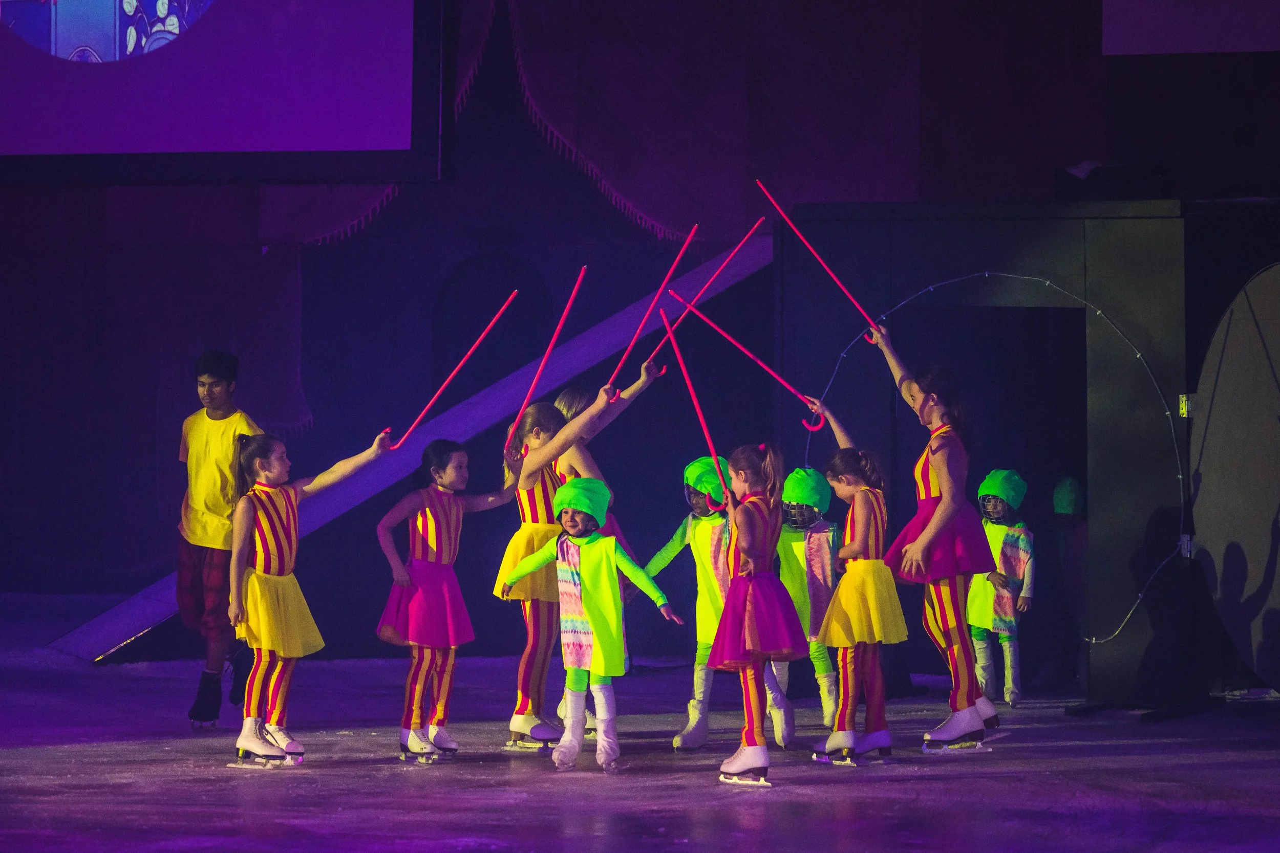 A group of performers on ice skates dressed in colorful, striped costumes with some wearing green hats and safety vests, holding hands and forming a circle, performing a choreographed routine under purple lighting.