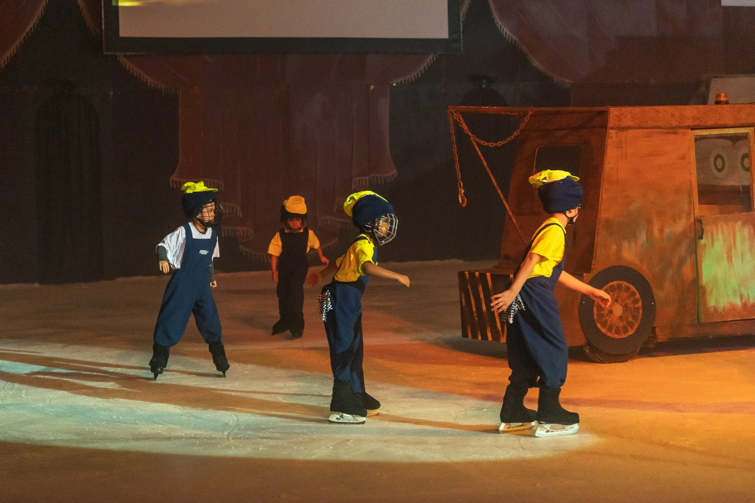 Children on roller skates practicing in an indoor skating rink with a vintage tractor prop in the background.