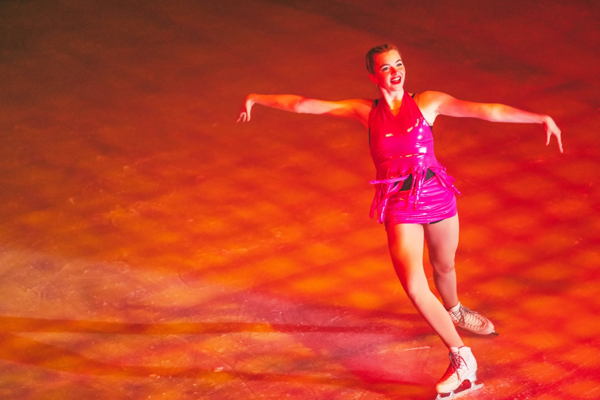 A young woman in a shiny red dress ice skating on a rink, with her arms outstretched and smiling.