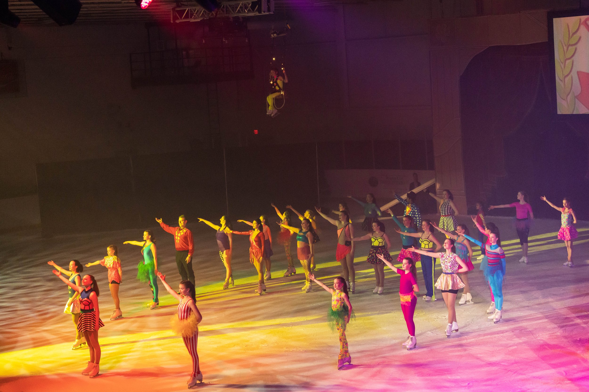 A large group of performers in colorful costumes on a stage, participating in a dance or musical performance, with some performers’ arms extended and a backdrop with lights.