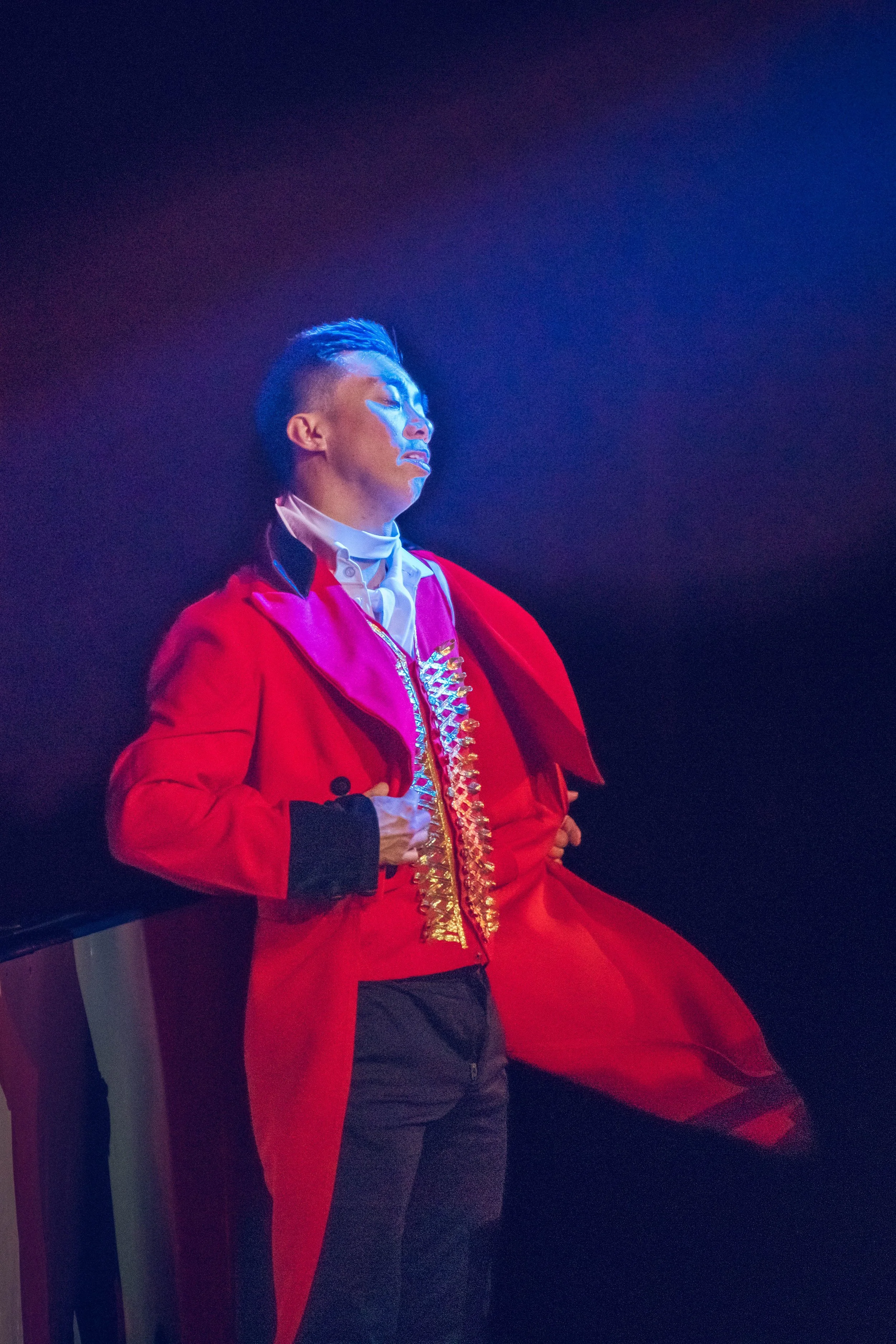 A person with short hair and light skin dressed in a red and gold costume with a cape, standing with eyes closed against a dark background, illuminated by blue light.