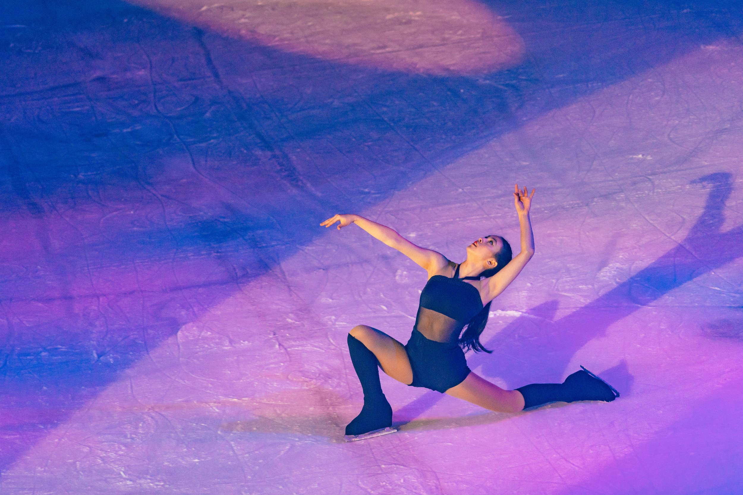 A female figure skater is performing a move on the ice, in a black dance outfit, with arms gracefully extended and one knee on the ice, against a backdrop of colorful purple and pink lighting.