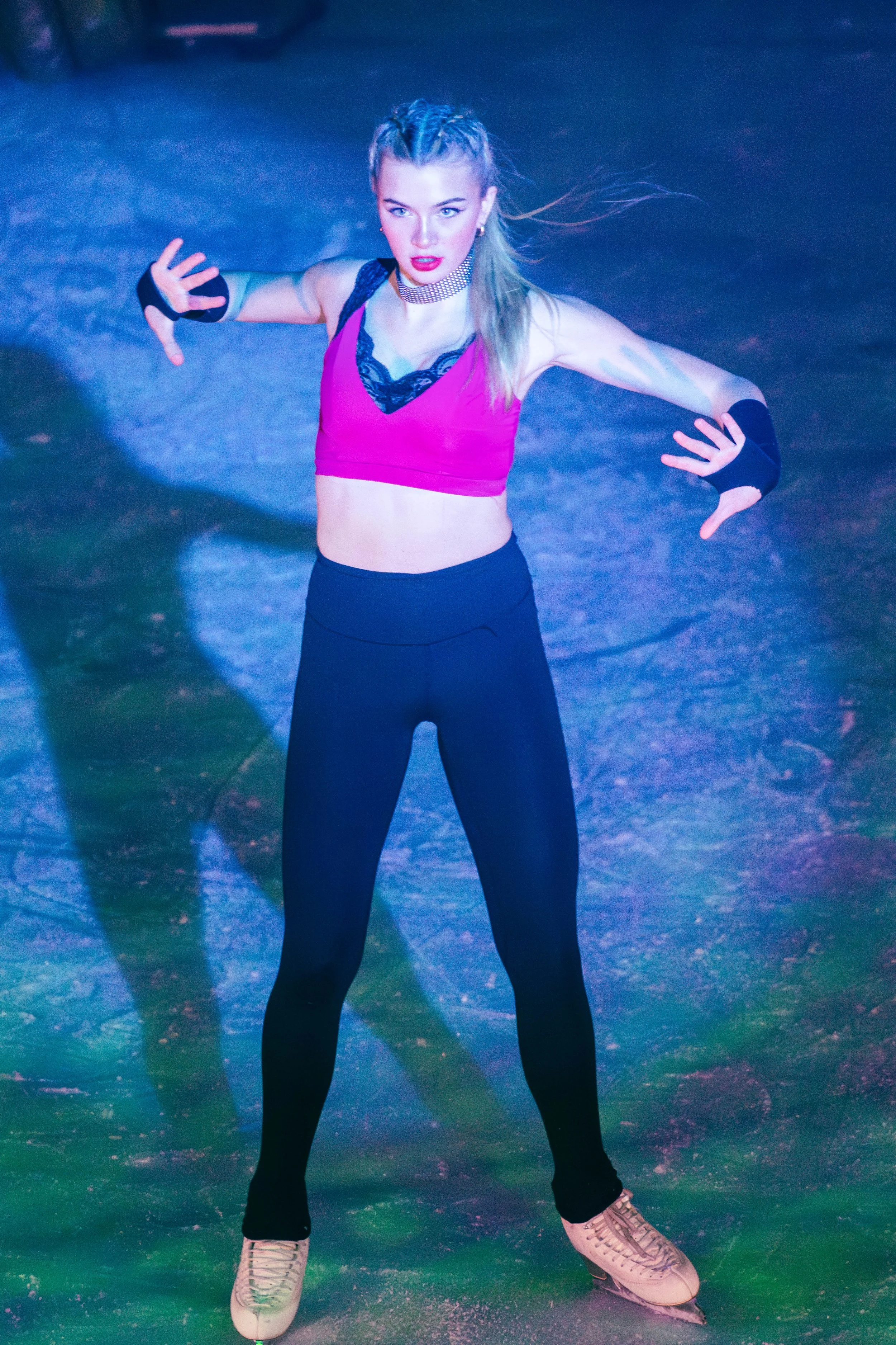 A young woman ice skating on an ice rink, wearing a pink sports top with black lace details, black leggings, white ice skates, and black gloves, with her hair styled in braids, striking a dynamic pose.