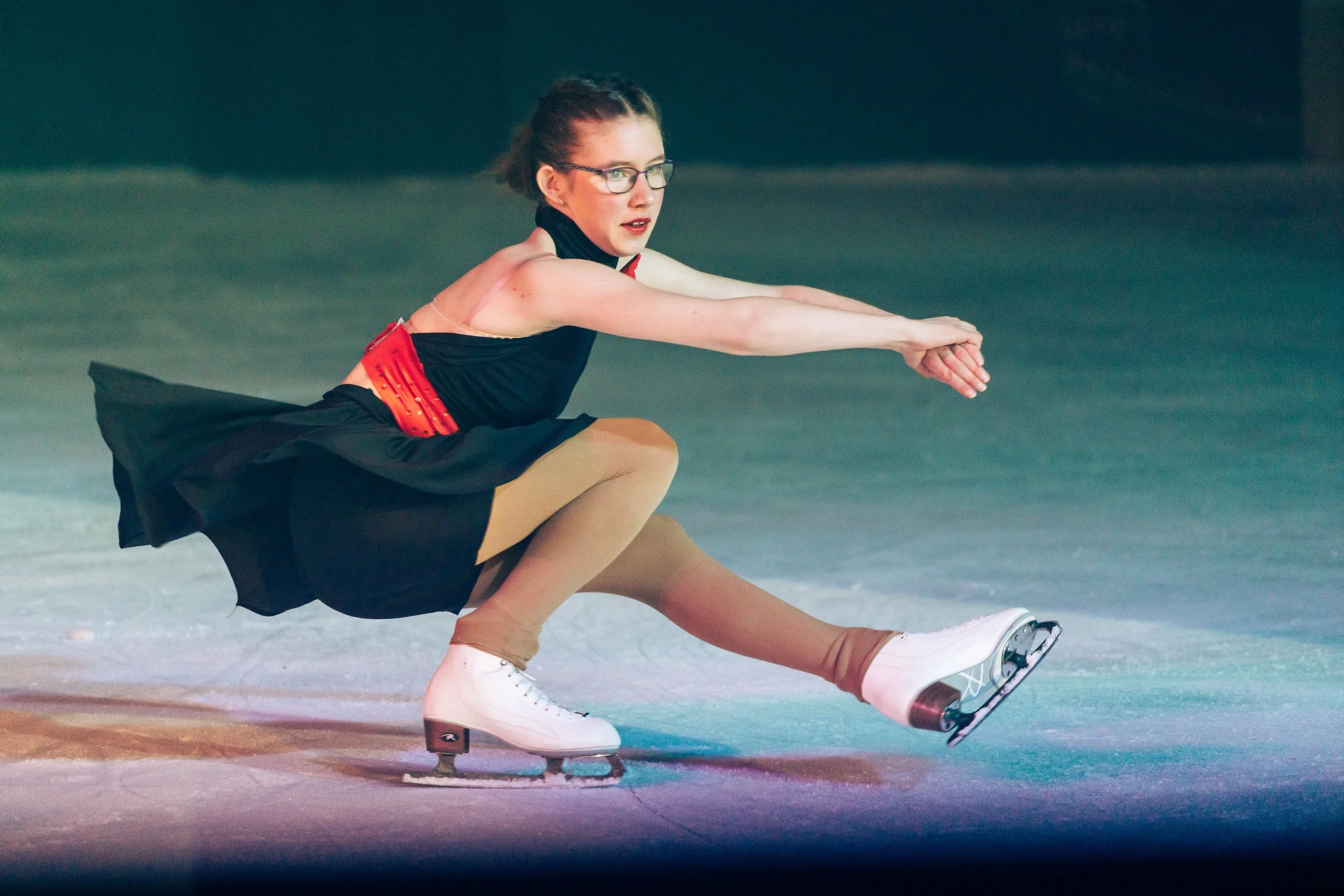 A female figure skater in a black dress with red accents performing a pose on the ice rink.