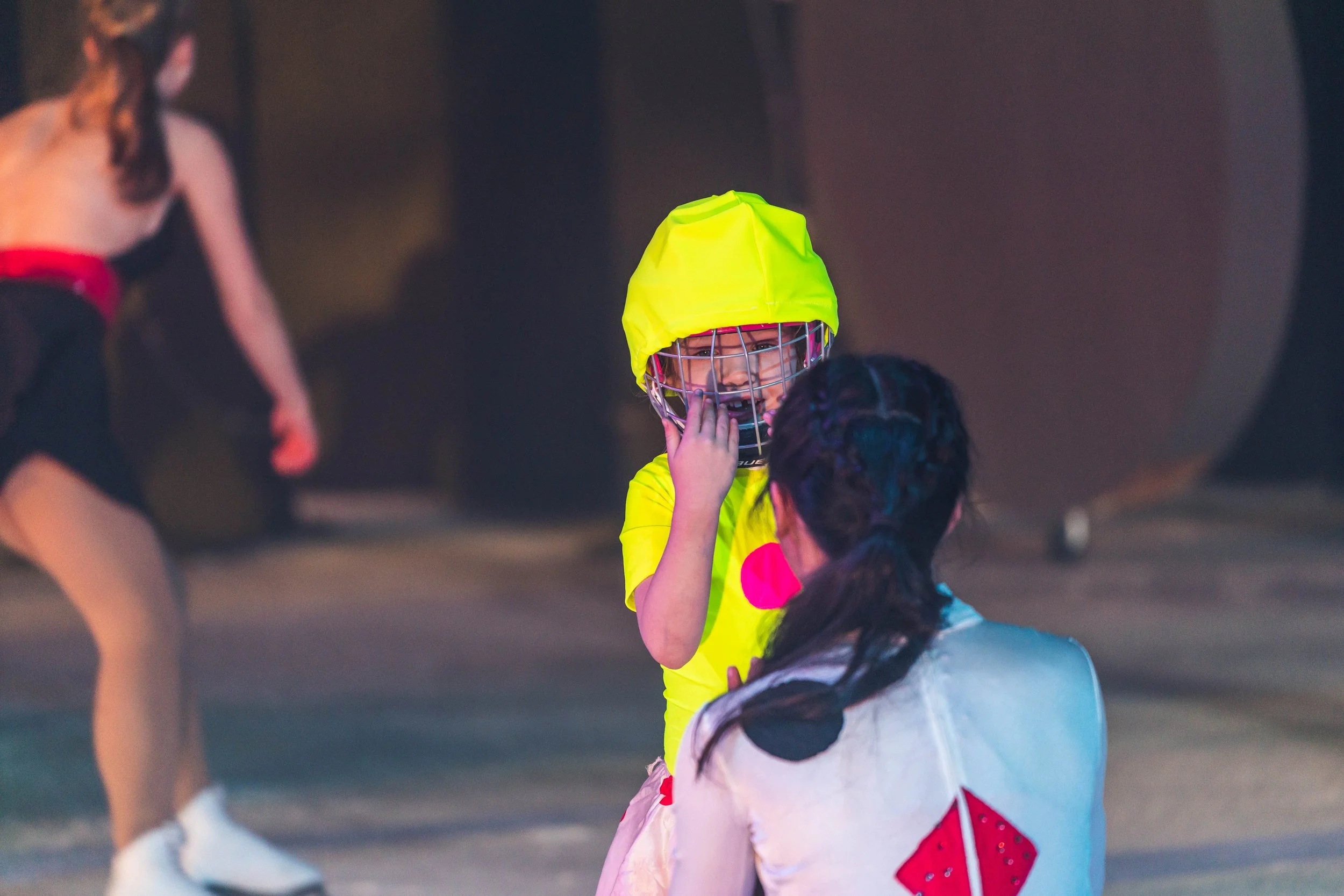A young girl wearing a yellow hockey helmet with a cage, yellow sports jersey, and pink gloves interacts with another girl wearing a white shirt with black spots and red accents, in an indoor setting with people in the background.