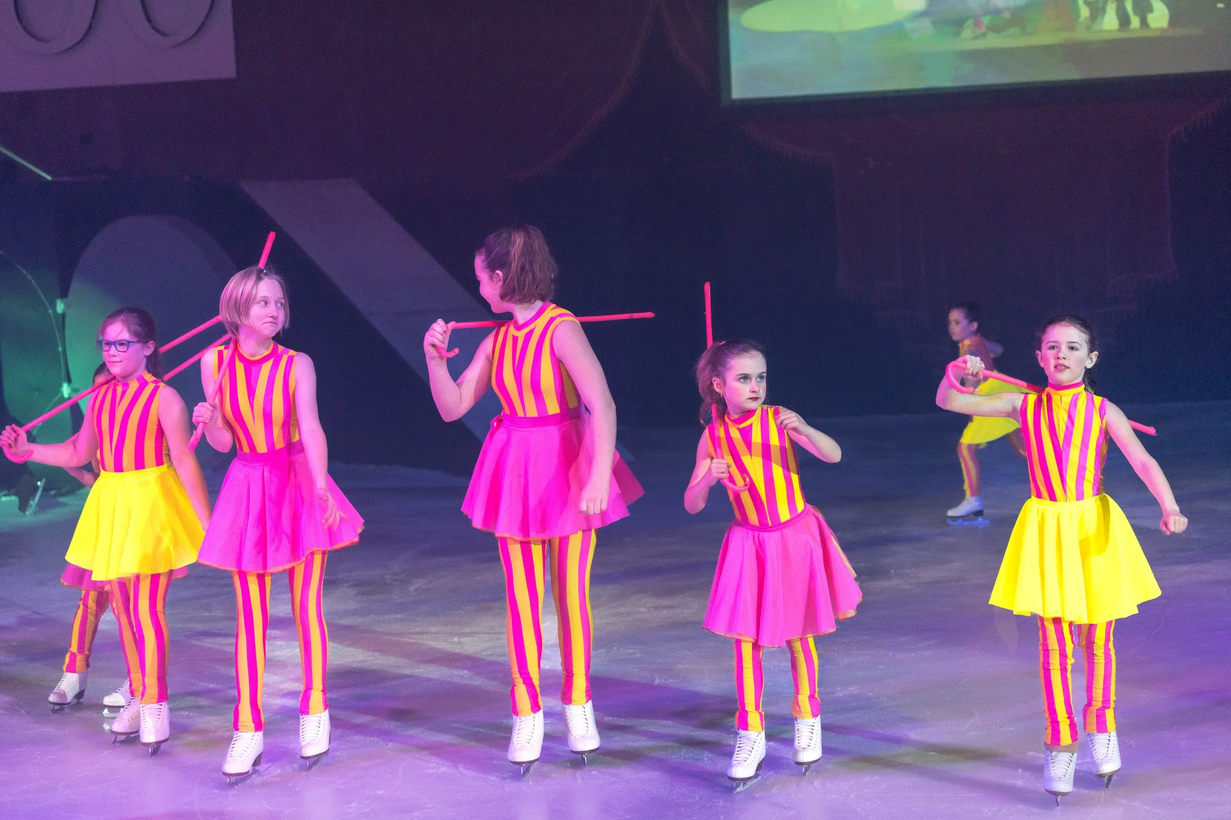 A group of young girls ice skating on an indoor rink, wearing colorful striped costumes in pink, yellow, and purple.