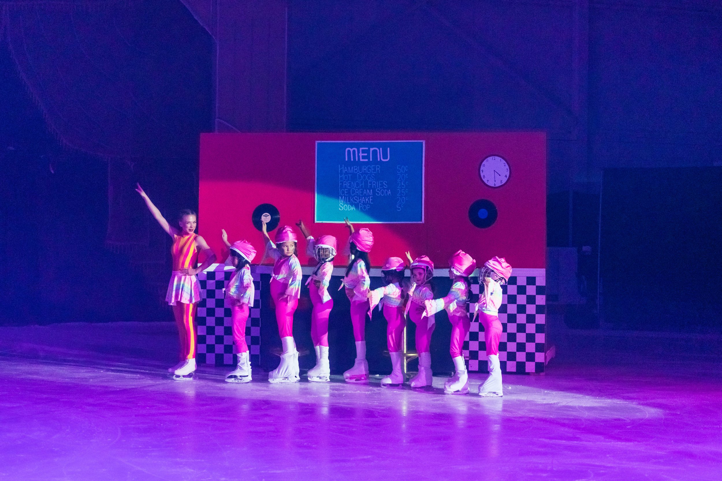 Children in pink costumes and helmets performing a dance on stage with a pink and black checkered backdrop, a large menu sign, and a clock.