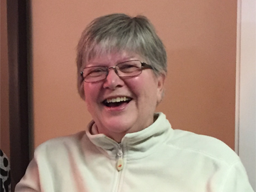 A woman with short gray hair and glasses smiling in a white zip-up jacket.