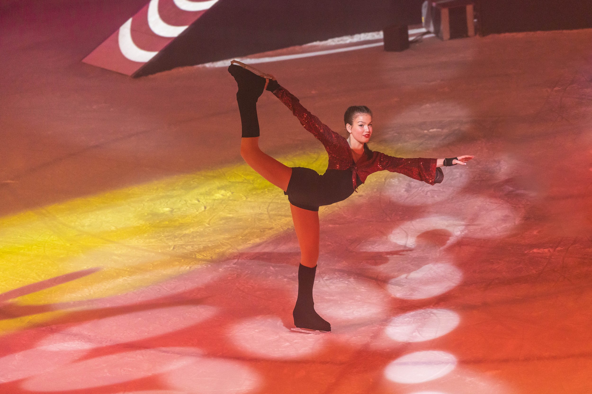 A female figure skater performing a Biellmann spin on the ice rink, wearing a red sequined top and black skirt.