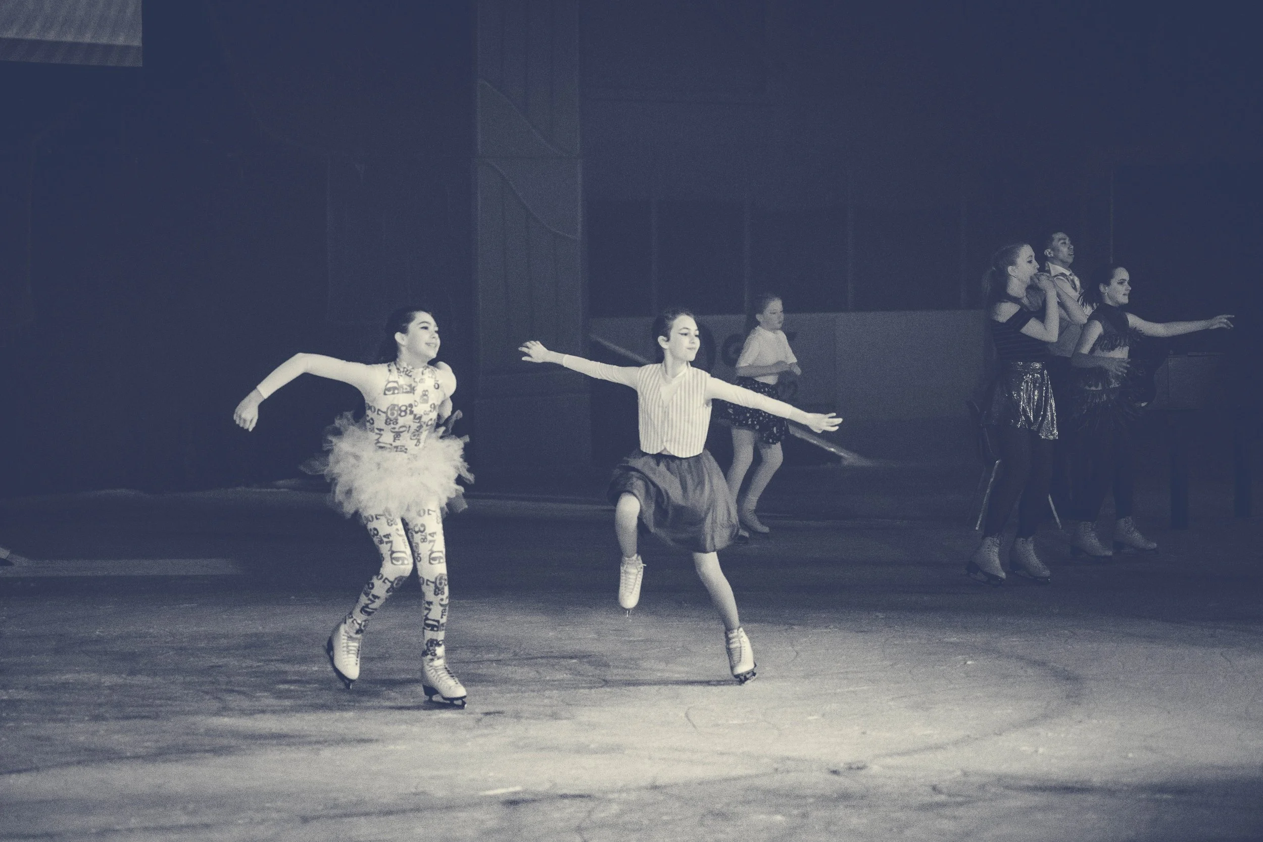 Group of young girls in dance costumes practicing ballet or jazz dance on stage, some with arms outstretched, others in different dance poses.