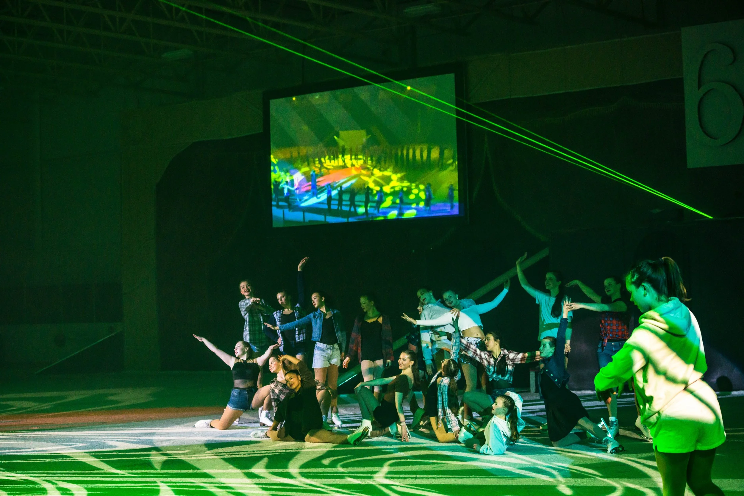 A group of young performers practicing dance on stage under green and yellow lighting with a large video screen in the background showing a dance scene.