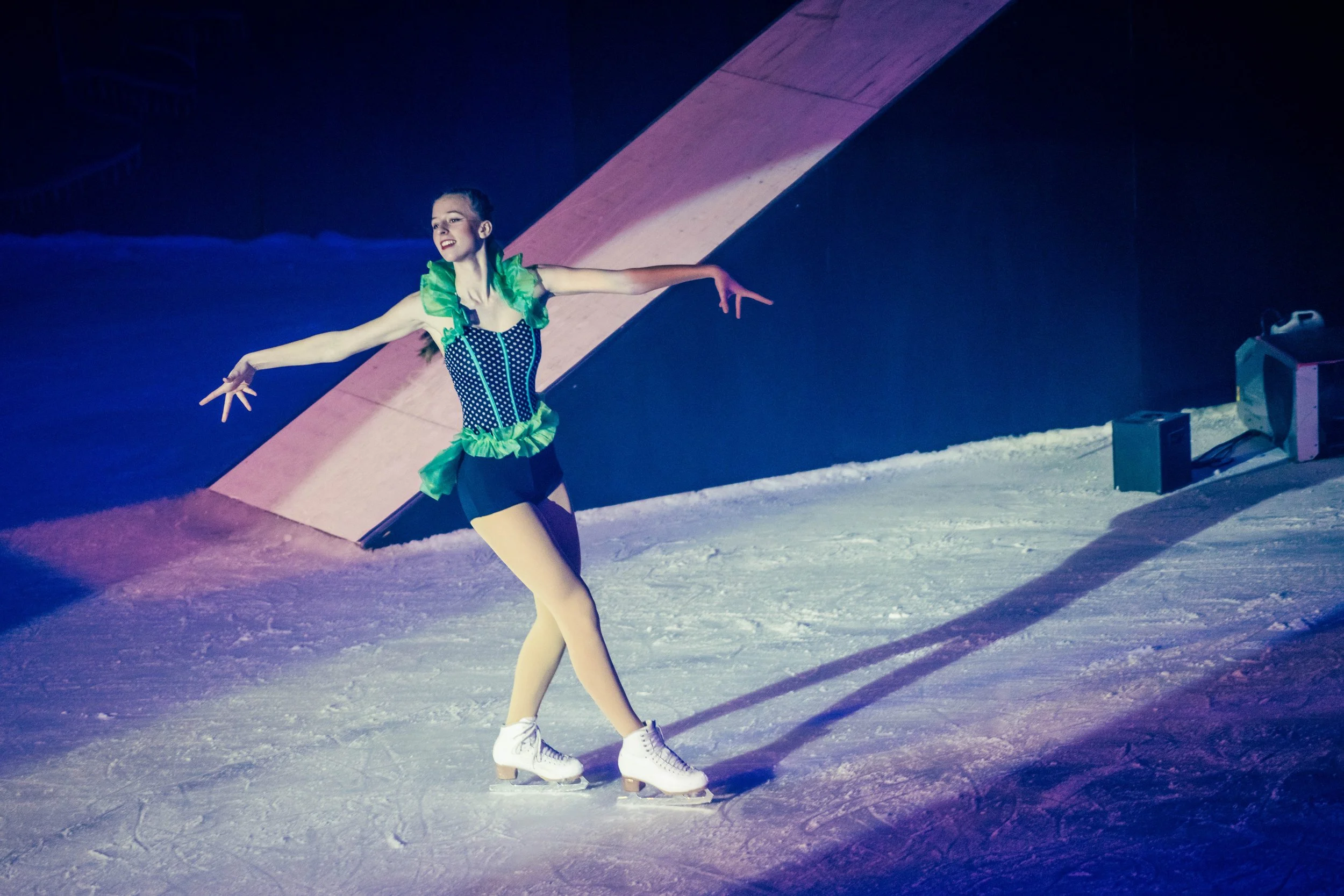 A female figure skater performing on ice in colorful costume, with arms outstretched and smiling, illuminated by stage lights.