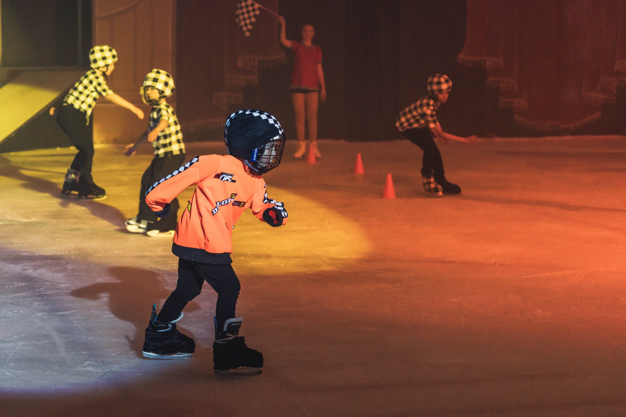 Children wearing helmets and skating on an indoor ice rink with cones, supervised by an adult in the background.