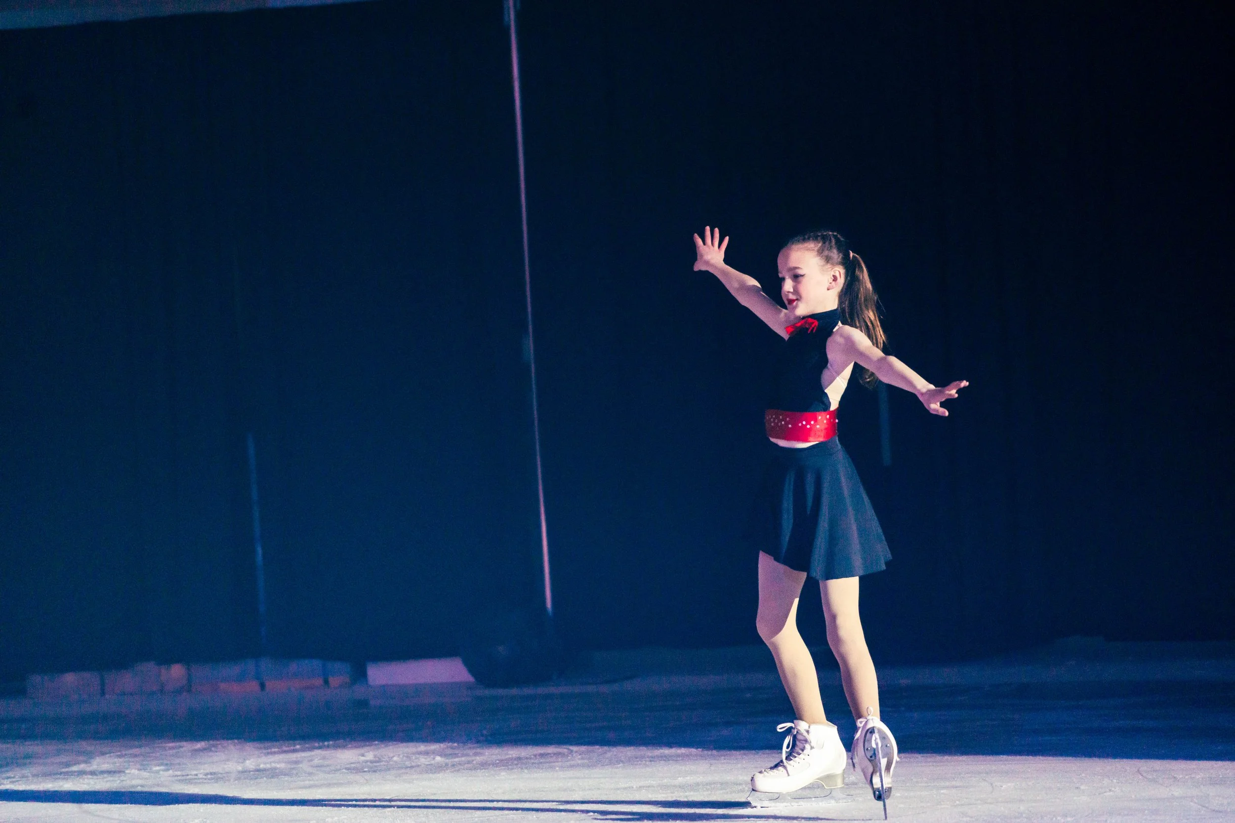 Young female figure skater in a black and red costume gliding across an ice rink indoors, extending her arms gracefully with a focus on her performance.