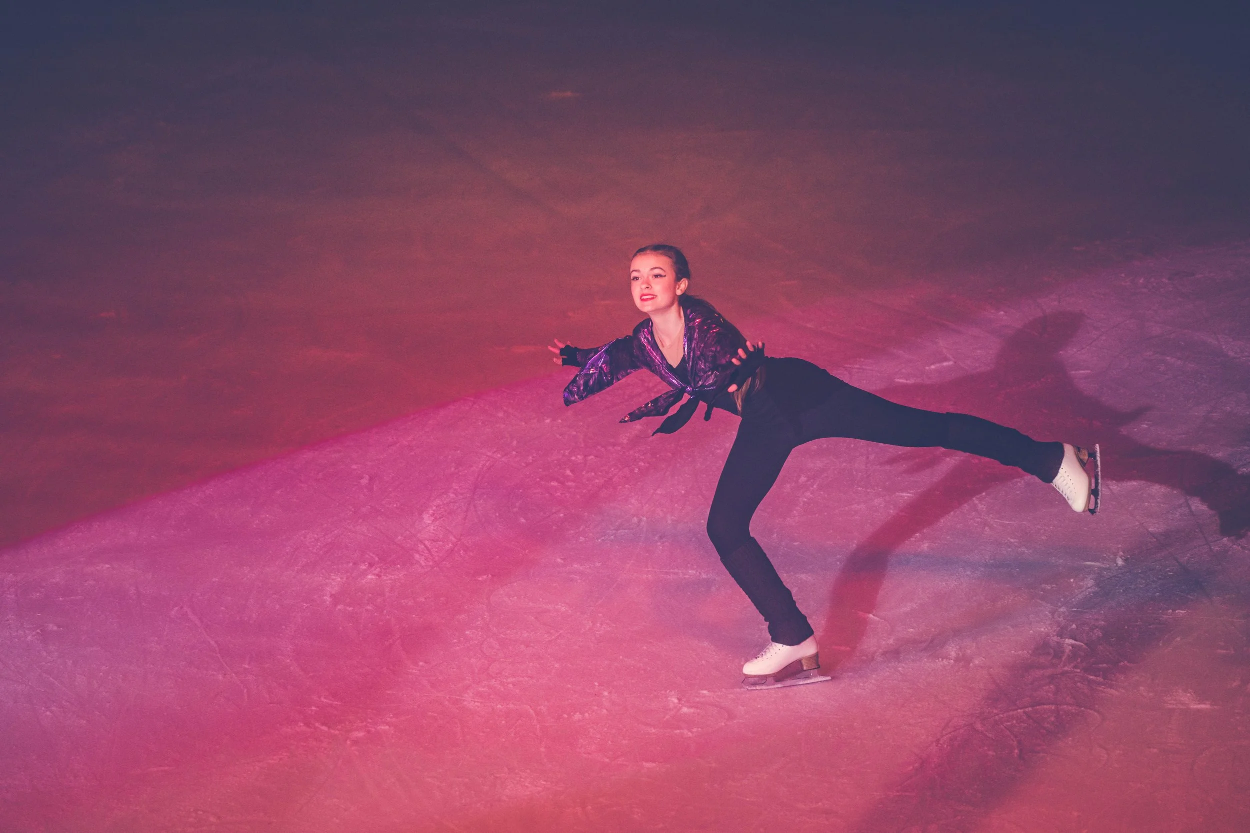 A young woman figure skater in a purple and black outfit performs a move on the ice rink. She is wearing white ice skates and a determined expression.