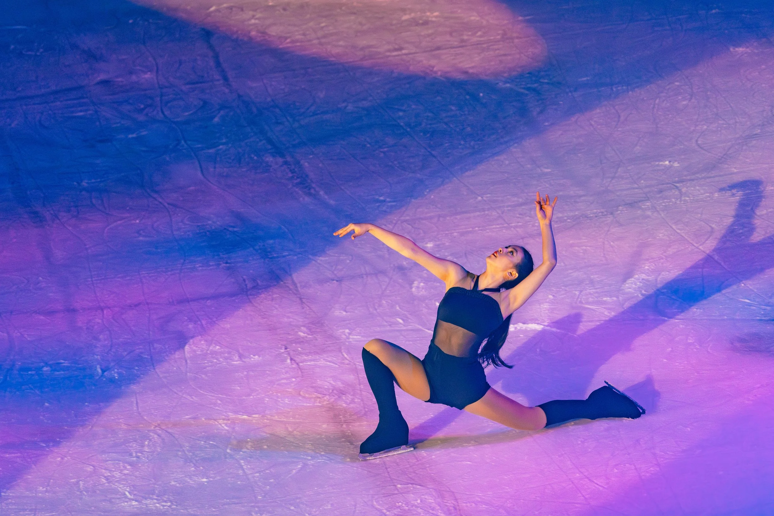 A female figure skater in black attire performing a pose on ice with purple and blue lighting creating a colorful background.