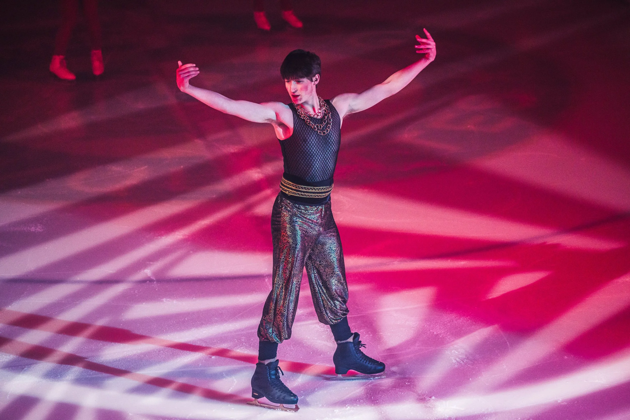 A male figure skater performing on ice, wearing a black mesh sleeveless top, patterned loose pants, black ice skates, and layered necklaces, against a background lit with pink and purple lighting.