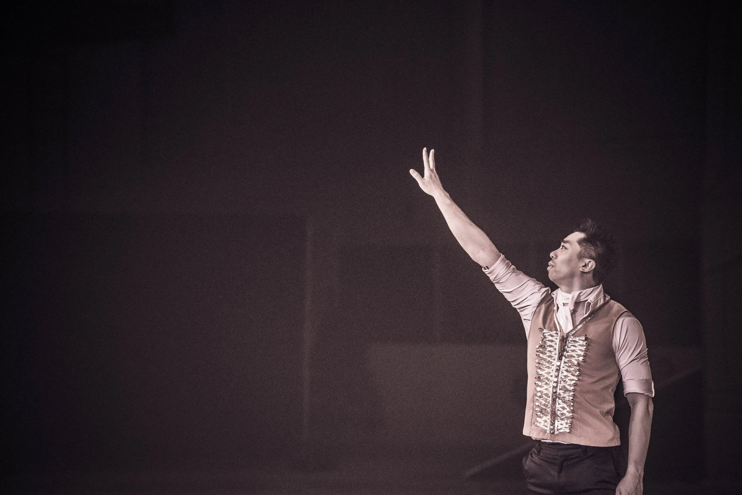 A man raising his right arm with two fingers extended, wearing a vest with shiny embellishments over a long-sleeve shirt, against a dark background.