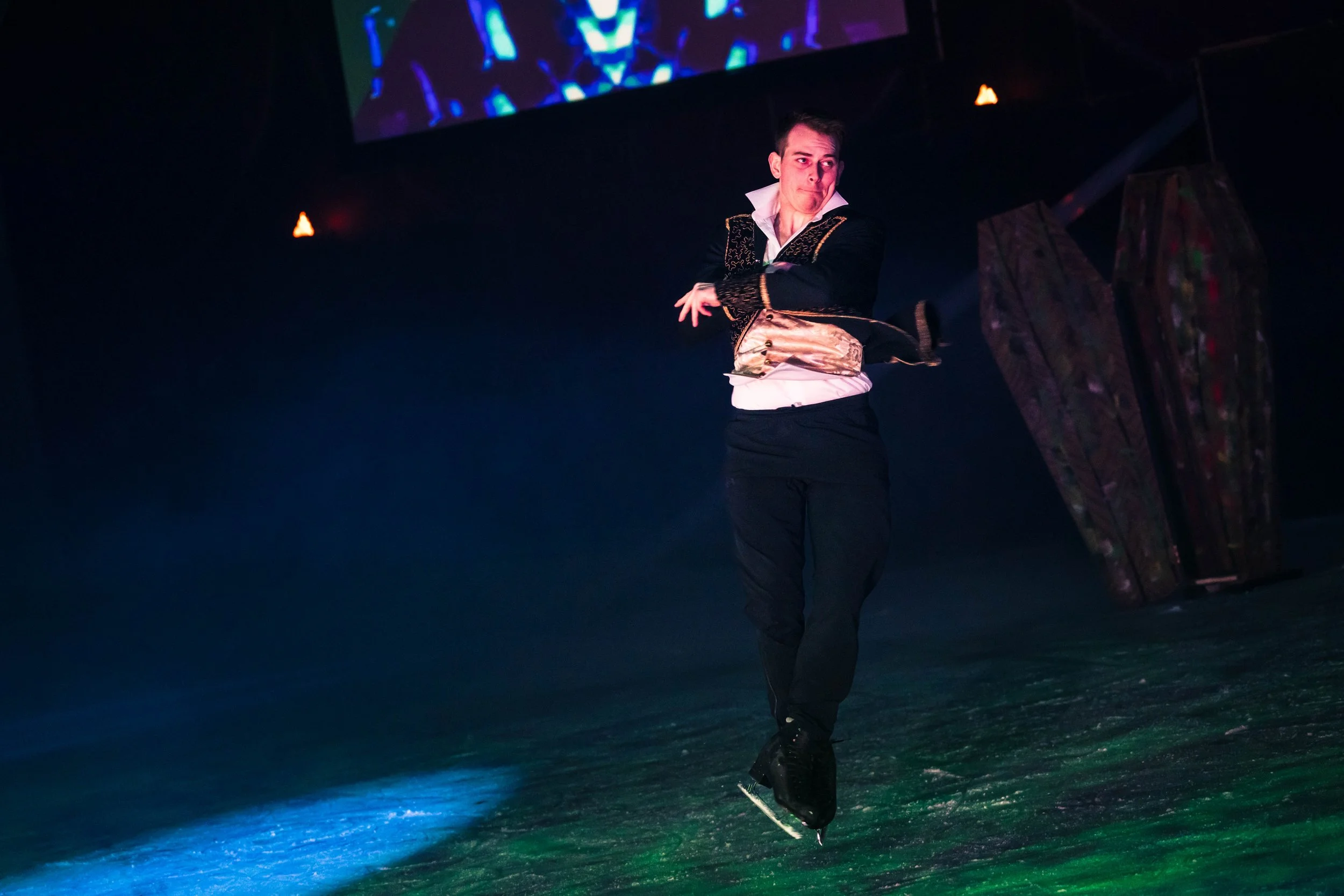 A male figure skater performing on ice with colorful lights reflecting on the surface, wearing a costume with gold and black details.